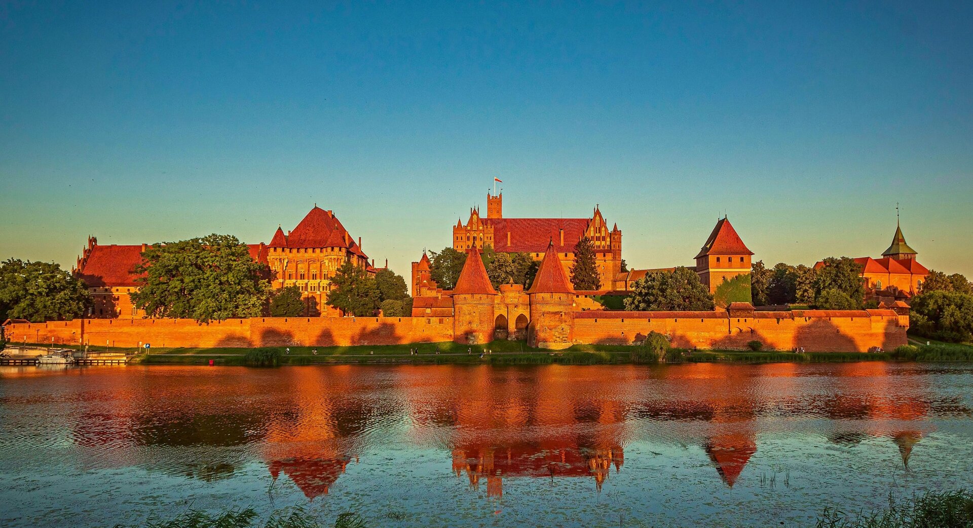 Zdjęcie przedstawia panoramę Zamku Krzyżackiego w Malborku. Zamek jest rozległy, otoczony murem. Stoi nad rzeką. 