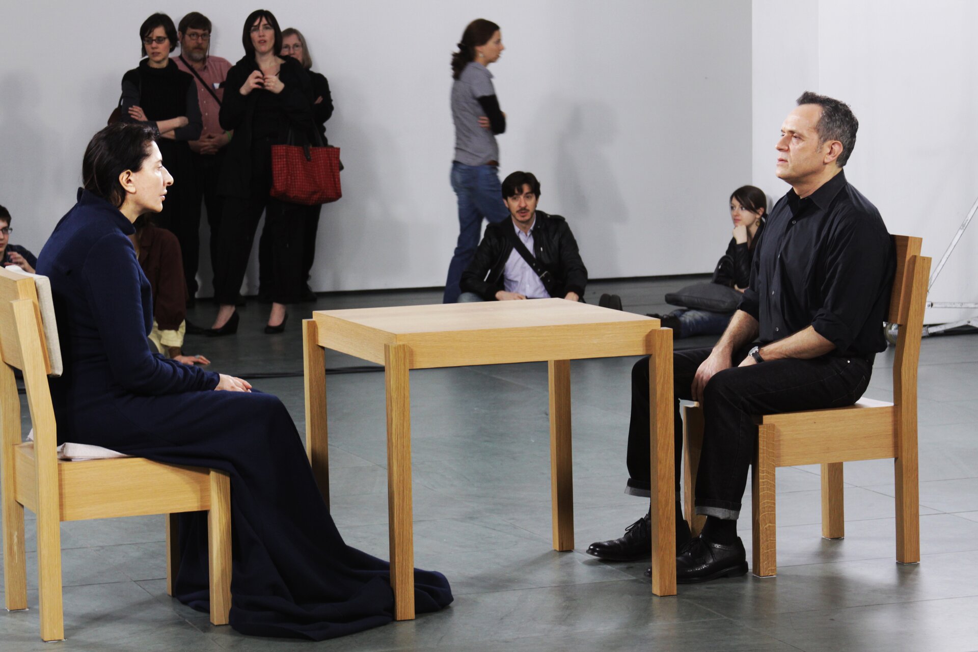 Zdjęcie przedstawia performance Mariny Abramowicz. W dużym pomieszczeniu stoi mały, drewniany stół. Siedzą przy nim, na przeciw siebie, kobieta i mężczyzna. Patrzą się na siebie. W ich tle widać obserwujących ich ludzi.