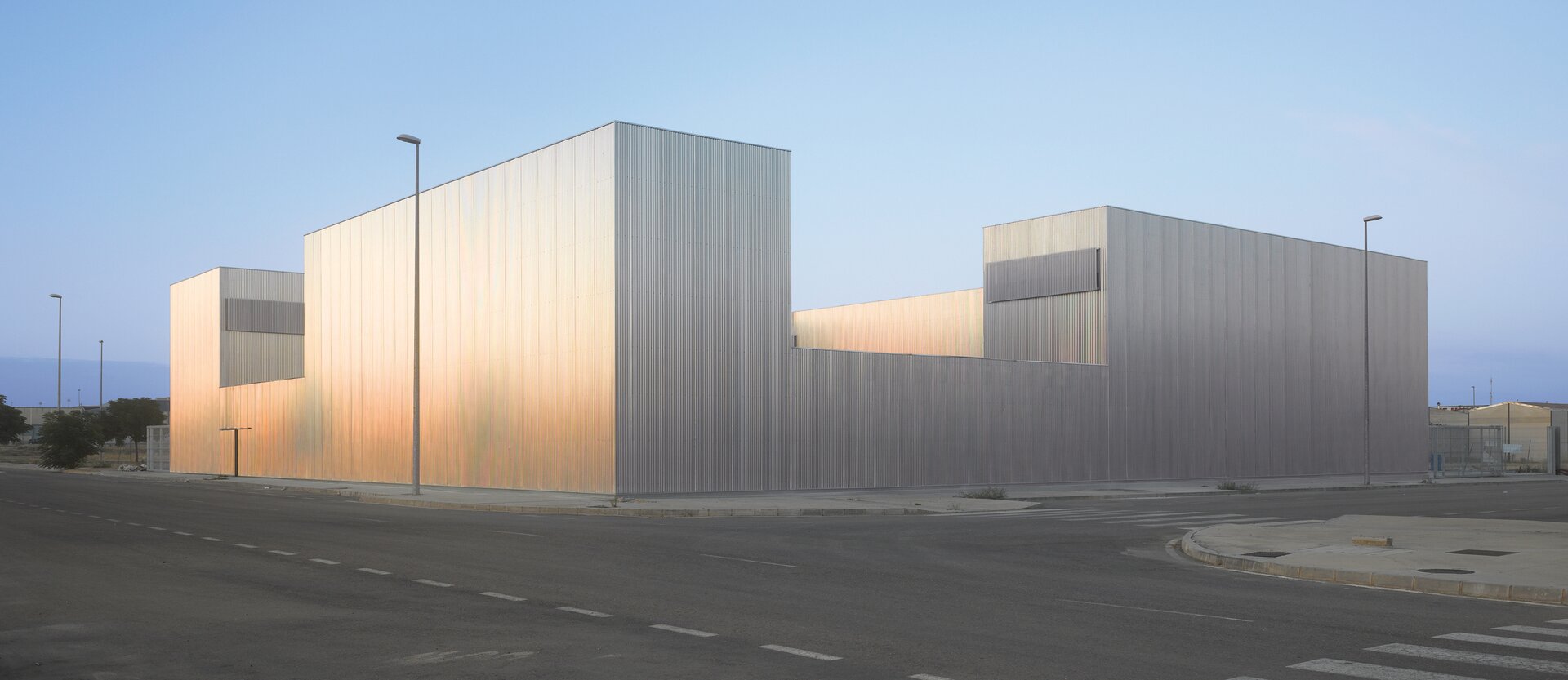 Fotografia przedstawia nowoczesny budynek, który stoi przy drodze. Budynek ma jasną elewację wykonaną z metalowych paneli w kolorze srebrnym. W budynku nie ma okien ani widocznych otworów wentylacyjnych. Z prawej strony budynku stoi ogrodzenie, a przed budynkiem lampy oświetlające chodniki.