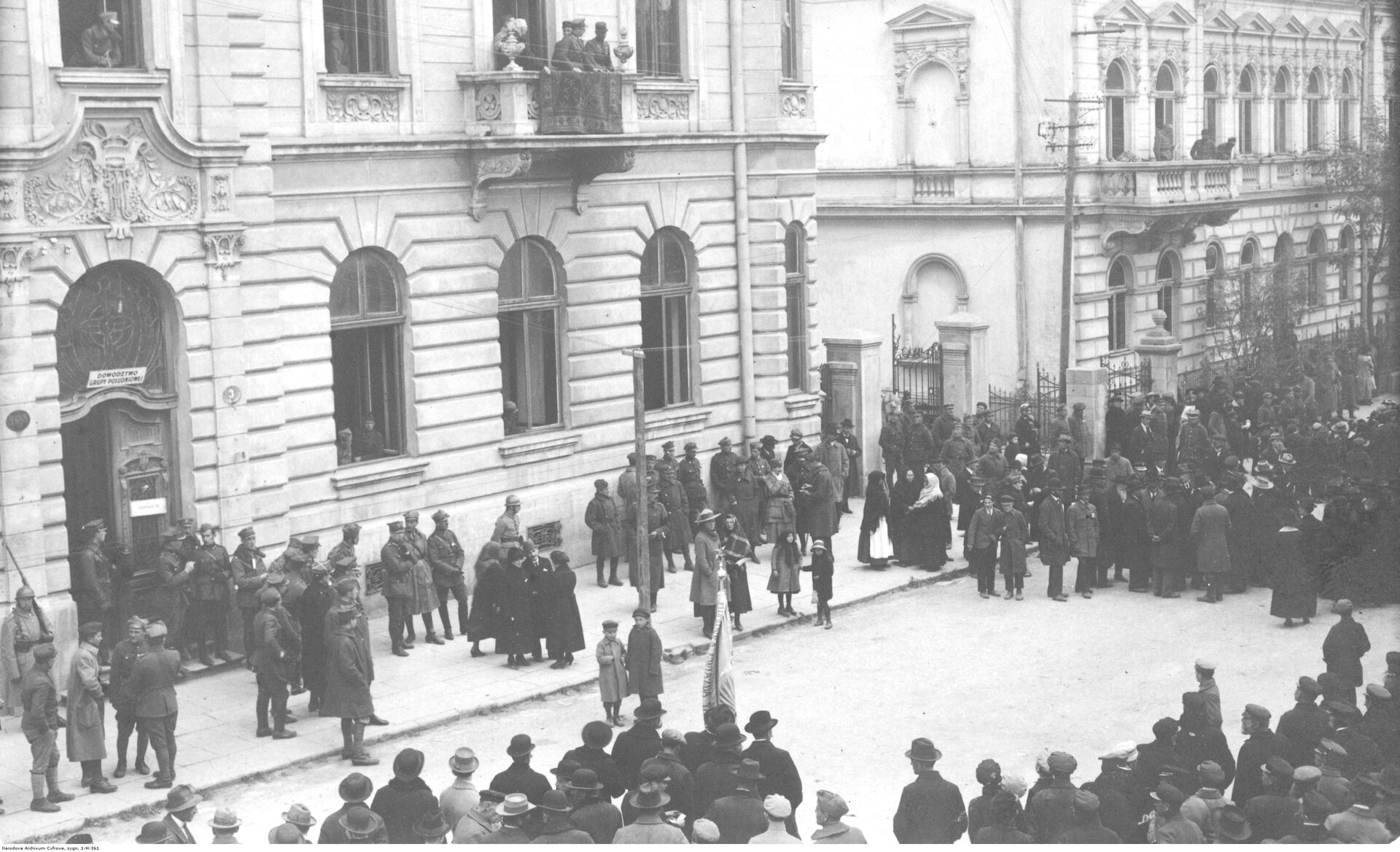 Fotografia przedstawia żołnierzy na ulicy jednego z miast. Widać też budynki mieszkalne i mieszkańców przyglądających się.
