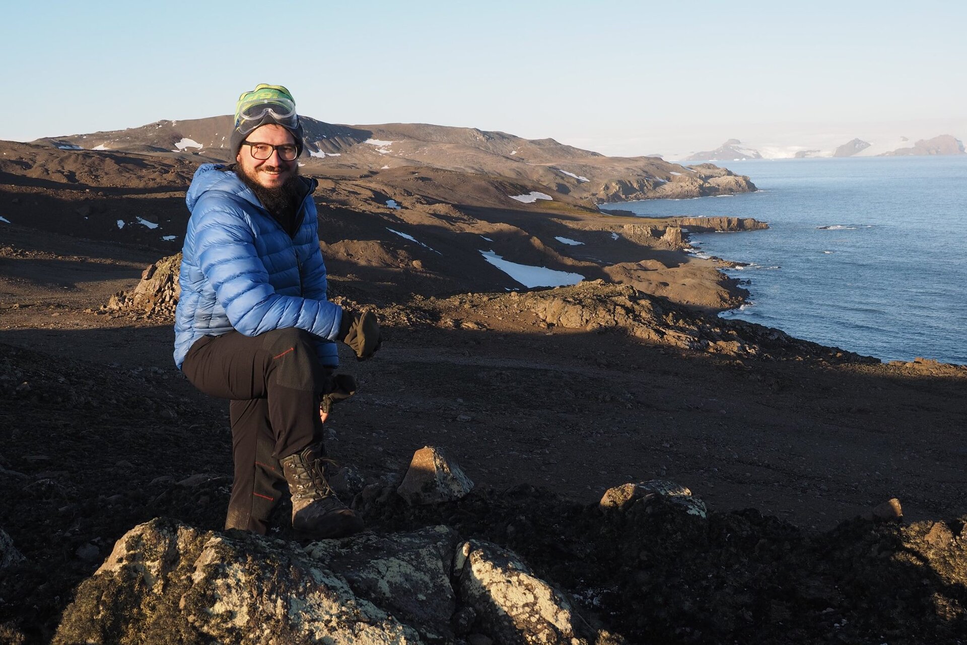 Fotografia badacza na tle wybrzeża w rejonie polarnym. Jałowy teren, ciemnoszare skały lub piasek. Na drugim planie wzniesienia i morze.