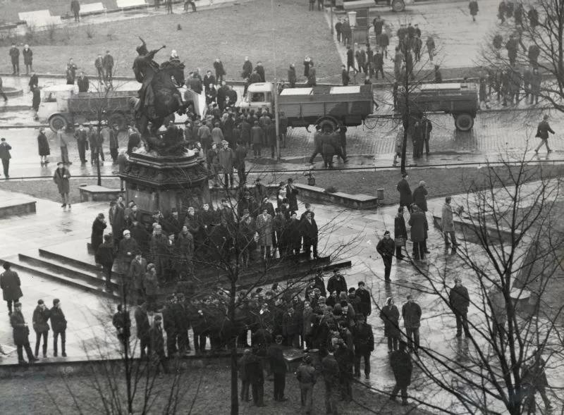 Zdjęcie przedstawia duży plac, na którym przy pomniku gromadzą się ludzie. Na ulicach otaczających plac stoją ciężarówki.