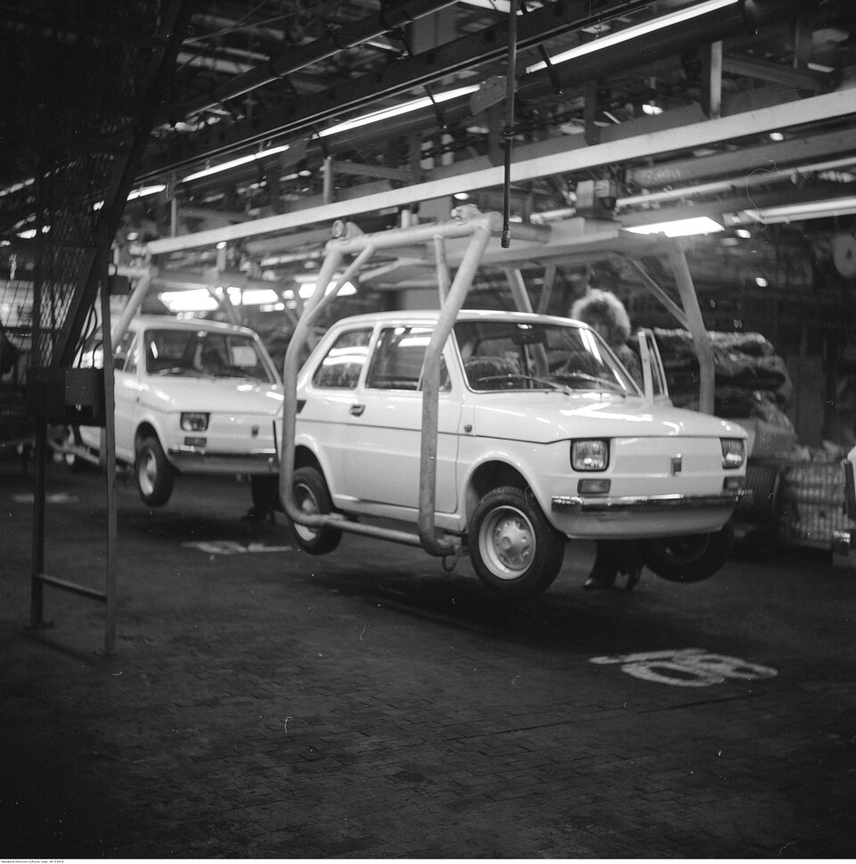 Zdjęcie przedstawia halę produkcyjną samochodu marki fiat 126p. Na pierwszym planie widoczne są podwieszone do sufitu samochody. 