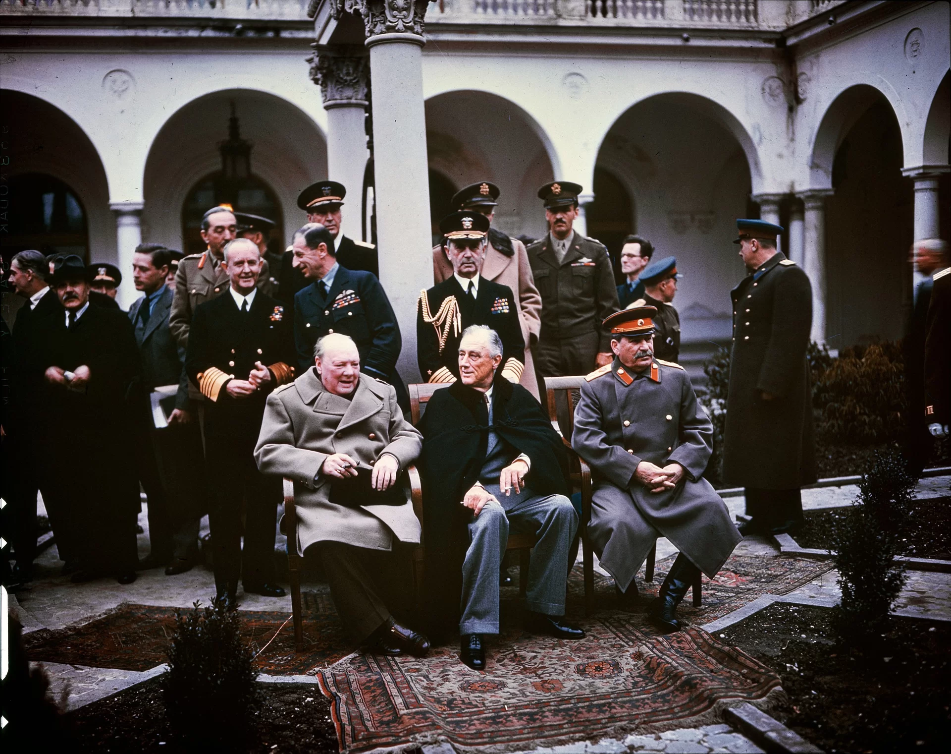Zdjęcie przedstawia Churchilla, Roosevelta i Stalina, którzy siedzą obok siebie na krzesłach. Churchill jest starszym, łysiejącym mężczyzną, ubrany jest w jasny płaszcz. Roosevelt również jest starszy, ma krótkie, siwe włosy oraz pociągłą twarz. Ubrany jest w garnitur, na który ma założony czarny płaszcz. W ręku trzyma papieros. Stalin ma siwe wąsy oraz szary mundur ze złotymi guzikami oraz czapką z czerwonymi elementami. Za mężczyznami stoją mężczyźni w mundurach i garniturach.