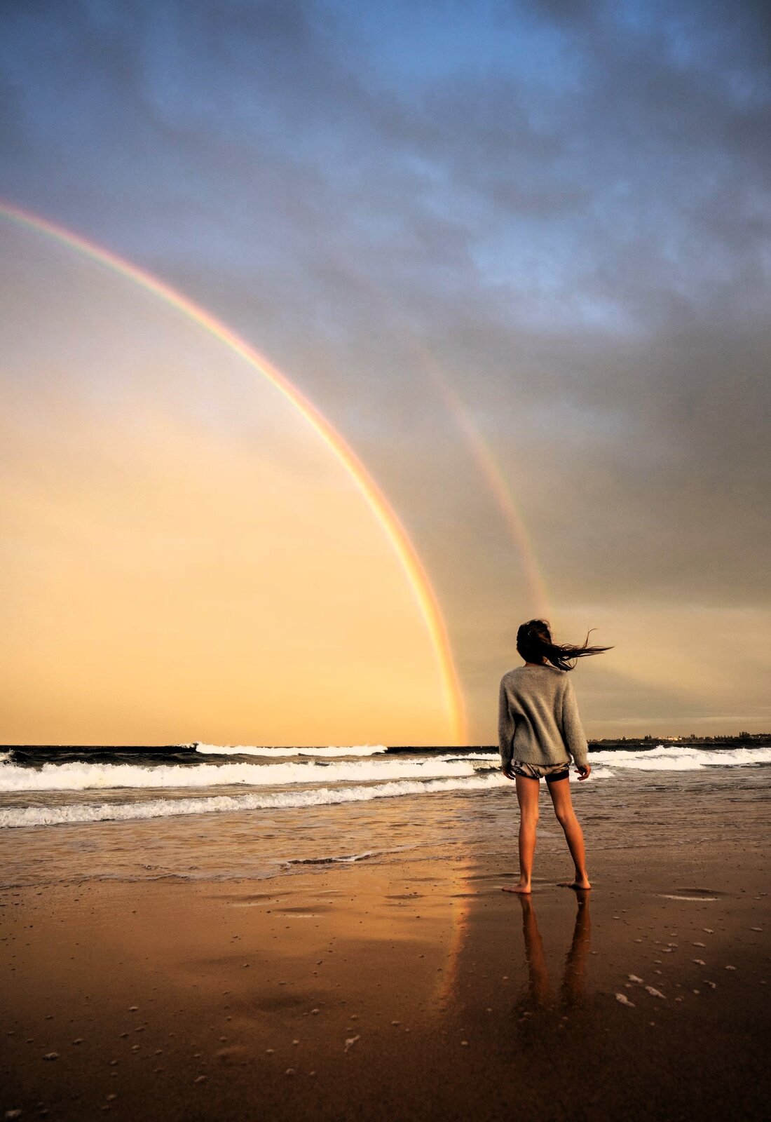 Zdjęcie przedstawia dziewczynkę stojącą na brzegu morza i patrzącą na tęczę. To chyba ciepłe popołudnie po deszczu. Morze niesie fale, a w mokrym piachu odbija się tęcza i postać dziewczynki w krótkich spodenkach, z rozwianymi włosami .
