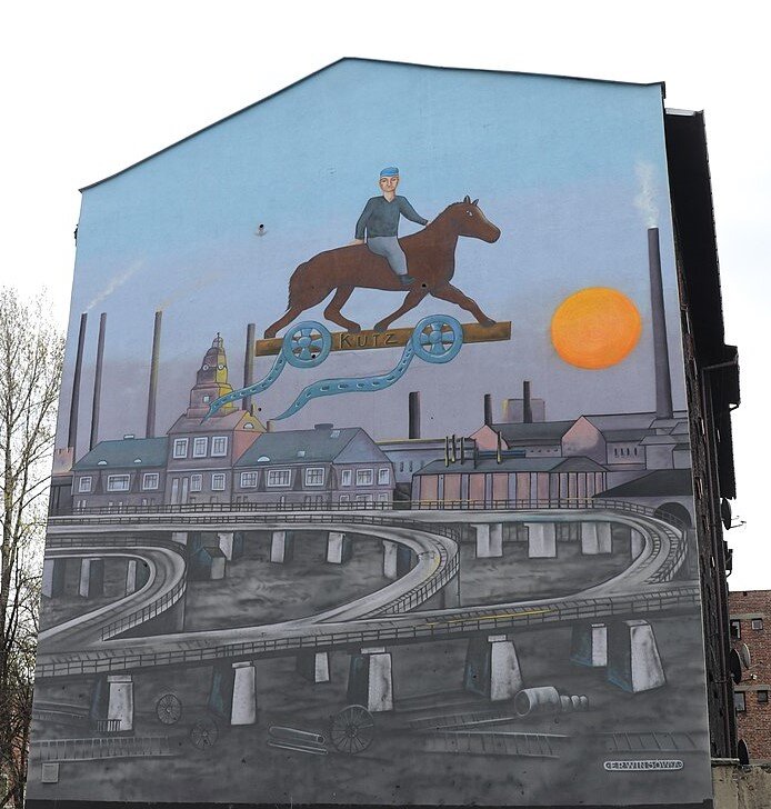 Zdjęcie przedstawia boczną ścianę wysokiej kamienicy. Jest na niej namalowany mural. Ukazana jest na nim rozwidlająca się droga oparta na przęsłach. Za nią znajdują się różne budynki fabryczne, liczne wysokie kominy. Nad całością, na niebie znajduje się mężczyzna jadący na koniu. Koń stoi na podstawie, której koła są rolkami. Rozwija się z nich taśma filmowa.