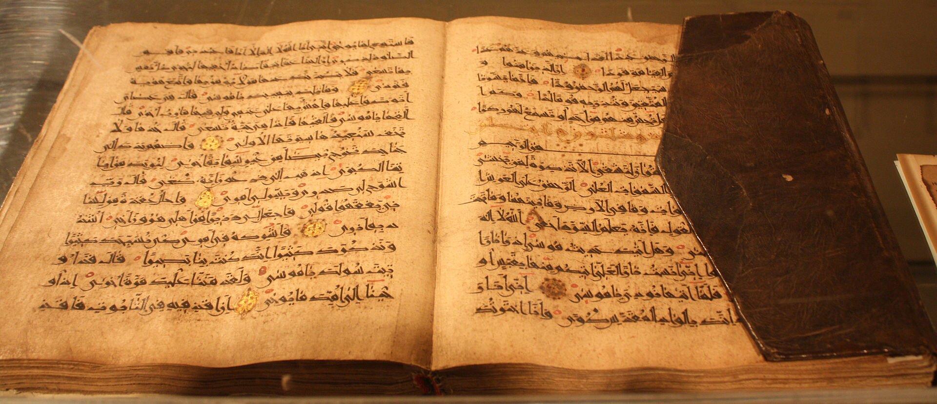 Ilustracja przedstawia otwartą książkę w czarnej oprawie zapisaną pismem arabskim. Zdjęcie jest w barwach pomarańczowo-brązowych.