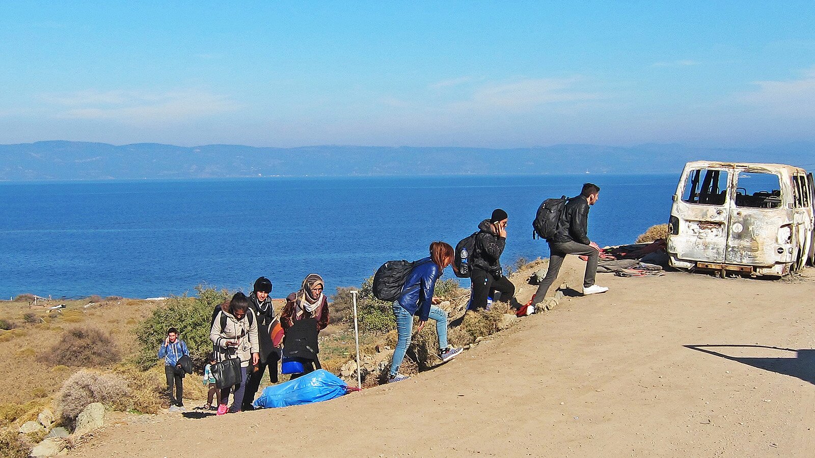 Zdjęcie przedstawia grupę ludzi z plecakami oraz torbami wchodzących po stromym zboczu na drogę. W oddali widoczne morze. 