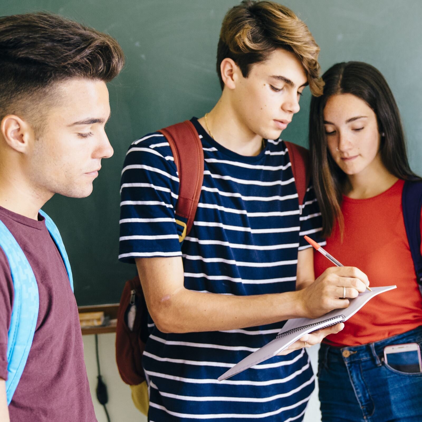 Zdjęcie przedstawia trójkę młodych ludzi stojących obok siebie. Po lewej stronie widoczny jest chłopak, w ujęciu od pasa w górę, zwrócony w prawo. Chłopak ma ciemne, krótkie włosy, jest ubrany w fioletową koszulkę z krótkim rękawem, na plecach ma niebieski plecak. Chłopak stojący w środku ma krótkie, jasne włosy, jest ubrany w koszulkę z krótkim rękawem w granatowe i białe paski, na plecach ma czerwony plecak. W lewej dłoni trzyma zeszyt, a w prawej dłoni długopis, którym coś notuje w zeszycie. Na prawo od niego widoczna jest dziewczyna z długimi, ciemnymi włosami, ubrana w czerwoną bluzkę i dżinsy. Dziewczyna ma pochyloną głowę o patrzy w kierunku zeszytu.