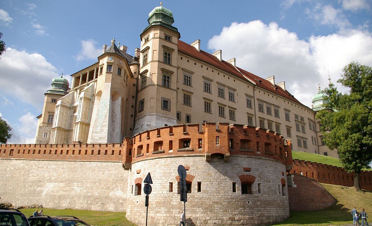 Fotografia barwna przedstawiająca Zamek Królewski na Wawelu od strony północno-wschodniej. W centrum zdjęcia znajduje się narożnik budynku z Wieżą Zygmunta III. Obok niej, po lewej - niższa wieża, zwana Kurzą Stopką. Zamek okala mur obronny. Pod Kurzą Stopką wybrzusza się,  przybierając  formę półokrągłej bastei - miejsca, z którego strzelano do wroga. Color photograph of the Wawel Royal Castle from the north-east. In the center of the photo there is a corner of the building with the Sigismund III Tower. Next to it, on the left - a lower tower, called the Kurza Foot. The castle is surrounded by a defensive wall. Under the Kurza Foot, it bulges, taking the form of a half-round bastion - the place from which the enemy was fired.