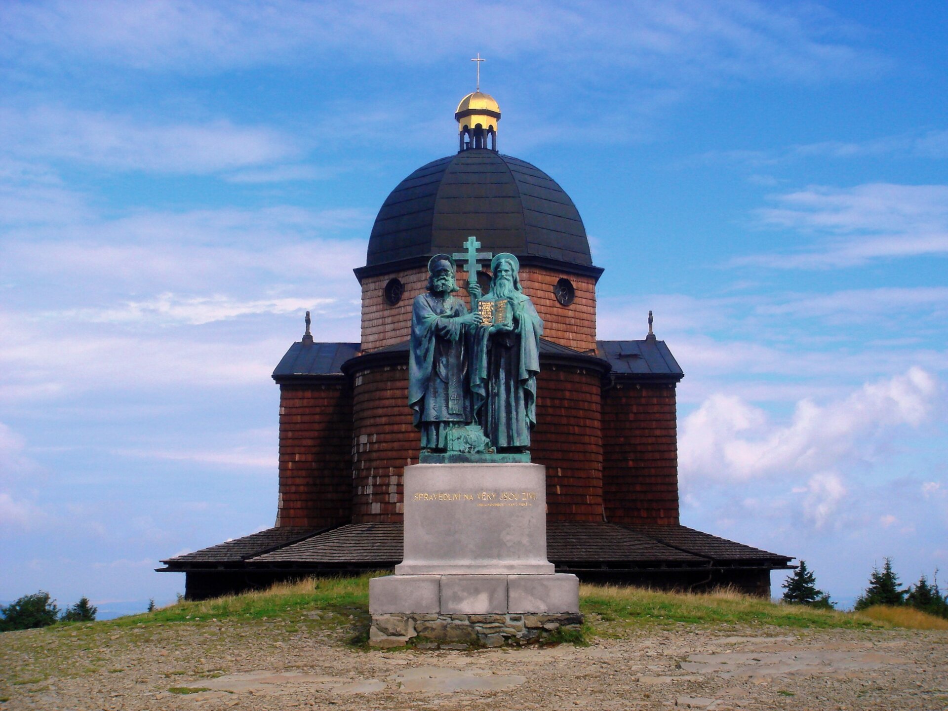 Widok współczesny na pomnik Cyryla i Metodego, ustawiony na kamiennym postumencie. Dwie figury dwóch mężczyzn w brodach patrzą na wprost widza. Podtrzymują stojący pomiędzy nimi krzyż obrządku wschodniego. Figura po prawej stronie to mężczyzna z dłuższą brodą i aureolą na głowie, w obu dłoniach na wysokości brzucha podtrzymuje otwartą księgę zapisaną alfabetem greckim. Stojąca figura po lewej stronie przedstawia mężczyznę z krótszą brodą i aureolą, którego prawa dłoń jest oparta o kartę księgi. Napisy na księdze wykonano ze złota. Na postumencie ledwo widoczny złoty napis w języku czeskim. W tle widoczna świątynia chrześcijańska z kopułą, wykonana z cegły i drewna, na szczycie kopuły widoczny złoty krzyż chrześcijański. Tło nieba pogodne, z chmurami.  