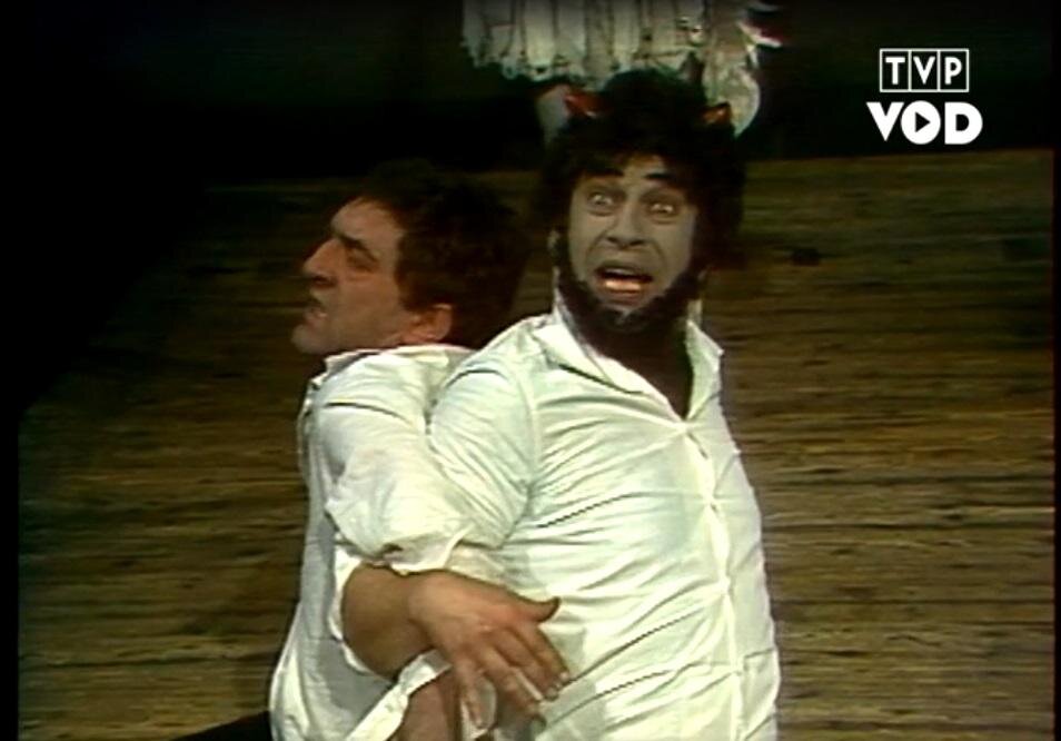 Ilustracja przedstawia dwóch mężczyzn w sile wieku. Postaci siedzą odwrócone do siebie plecami. Ręce mężczyzn są splecione na wysokości stawów łokciowych. Postać zaprezentowana po prawej stronie ukazuje owalną twarz w całości. Ma na głowie rogi i gęste, kręcone, ciemne włosy oraz brodę. Ma ciemne brwi, wytrzeszczone oczy i otwarte usta odsłaniające zęby. Drugi mężczyzna ukazuje lewy profil. Ma ciemne, krótkie włosy, gładkie czoło, ciemne brwi, haczykowaty nos i grymas niezadowolenia na twarzy. W tle ukazana jest drewniana ściana.