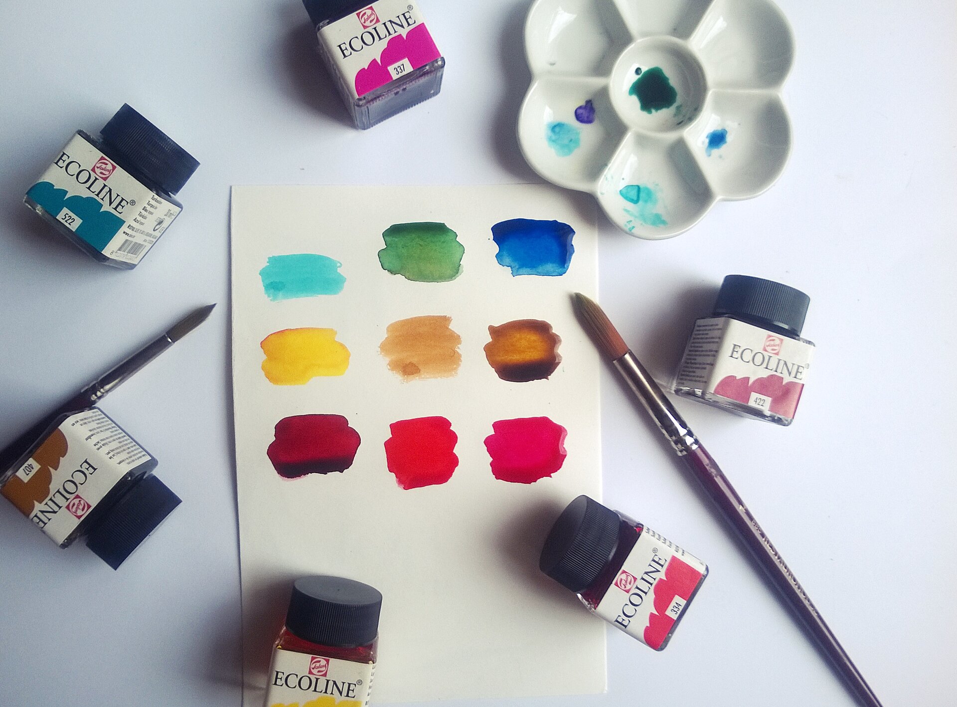 Ilustracja przedstawia akcesoria do malarstwa akwarelowego. W centrum zdjęcia, na białym tle znajduje się kartka z namalowanymi dziewięcioma kolorowymi plamami. Naokoło rozłożone jest sześć farb w szklanych pojemniczkach, dwa pędzle oraz mała, biała paleta do mieszania kolorów w kształcie kwiatu o sześciu płatkach.
