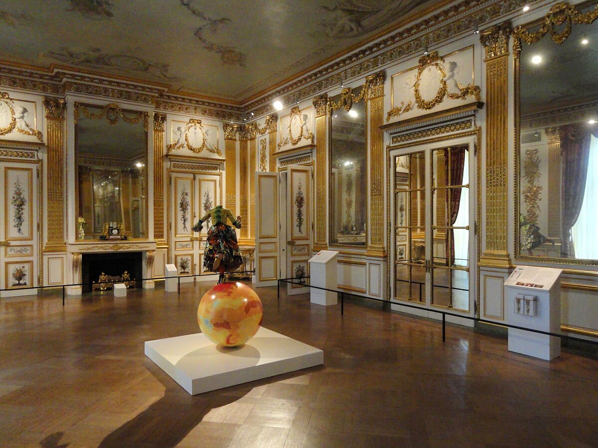 Fotografia barwna przedstawiająca Salon Doré - jedną z sal w Corcoran Gallery w Waszyngtonie. To jasna, obszerna sala ze ścianami bogato zdobionymi pozłacanym drewnem, pozłacanym brązem i antycznym alabastrem. Niektóre ze zdobień przyjmują kształt złotych pilastrów, czyli płaskich filarów, inne - złotych kwiatowych girland. Nad drzwiami medaliony z płaskorzeźbami amorków. Wielkie lustra ujęte w złote ramy. Sufit otoczony złotym gzymsem, podłoga wyłożona parkietem. Salon Doré, czyli Złoty Salon, został  zaprojektowany w 1770 roku przez Jeana Françoisa Chalgrina, francuskiego architekta. Pośrodku Salonu znajduje się instalacja współczesnego artysty Yinki Shonibare'a. Na kuli ziemskiej stojący na jednej nodze kobiecy bezgłowy manekin, ubrany w długą, wielobarwną suknię z falbanami. Druga noga uniesiona w tył. Stopy w kolorowych trzewikach. Ręce, lekko zgięte w łokciach, uniesione na poziomie klatki piersiowej. Praca nosi tytuł: Girl on Glob 2.