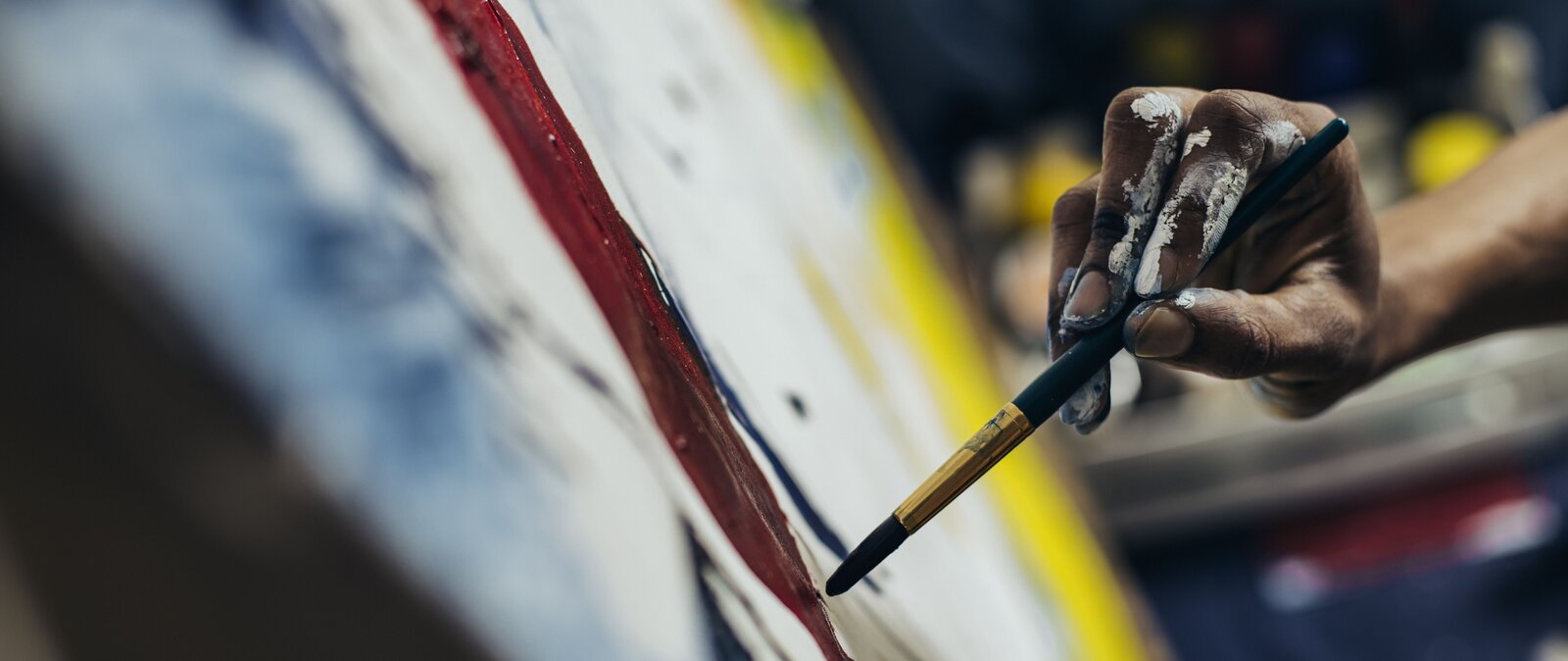 Kliknij, aby powiększyć. Panoramiczna fotografia barwna w układzie poziomym przedstawiająca w zbliżeniu dłoń artysty malarza przy pracy. Tylko część fotografii ma zachowaną ostrość obrazu. Tak ukazane zostały: wyłaniająca się zza prawej krawędzi kadru dłoń na pierwszym planie, brązowa plama farby i trzymany tuż nad malowidłem niewielki pędzel. Jest on okrągły, z czarnym włosiem i trzonkiem. Metalowa skuwka pędzla ma barwę złotą. Palce, które go przytrzymują noszą liczne ślady białej farby. Powstający obraz przecina kadr skosem – na środku i po lewej stronie. Ciemne i niebieskie plamy w dolnym rogu na pierwszym planie oraz biel podłoża i żółte plamy w głębi kadru, na odległym krańcu obrazu, są nieostre. Podobnie, znajdujący się jeszcze dalej w tle przysłonięty dłonią malarza blat stołu z kolorowymi słoikami farb. Nie jest on ukazany w poziomie. Opada na lewa stronę, co wskazuje na to, że fotograf, by dodać dynamiki zdjęciu, przechylił nieco aparat od pionu w trakcie wykonywania fotografii.