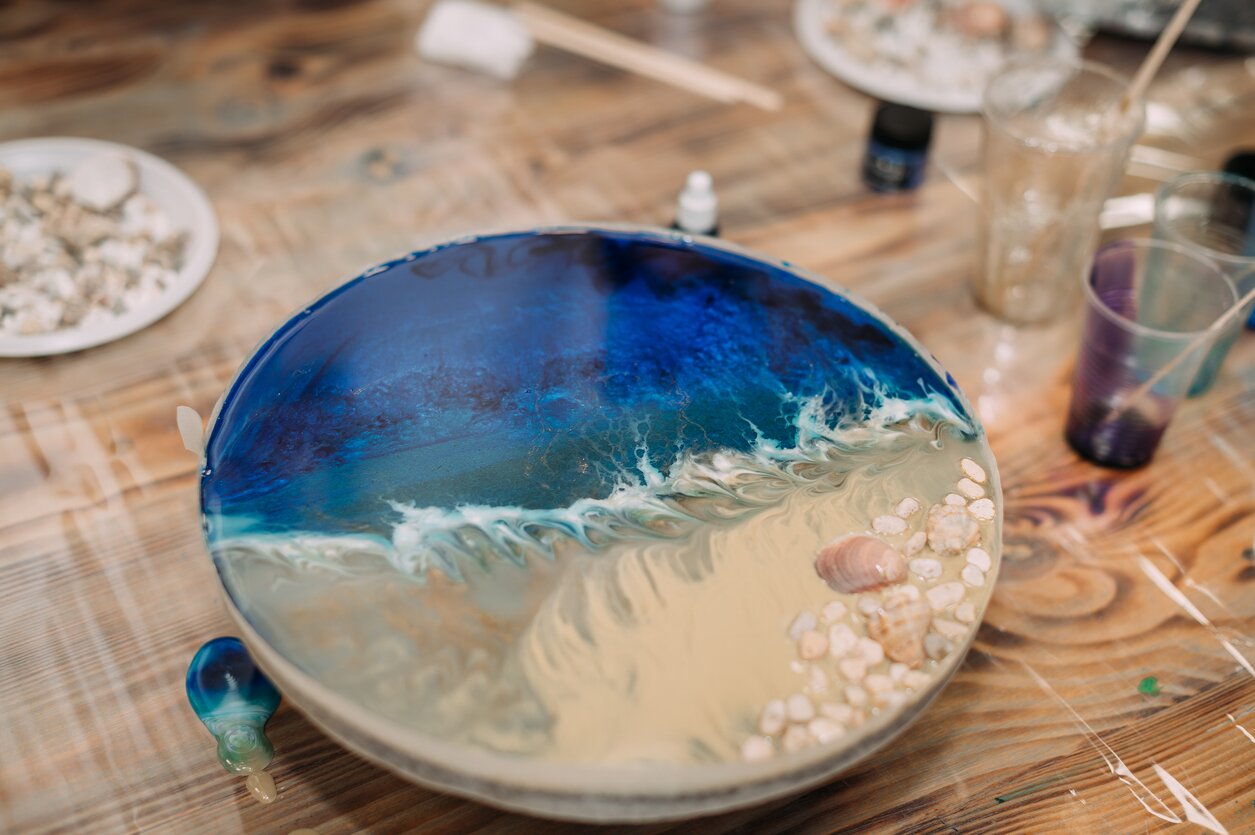 Zdjęcie przedstawia element architektoniczny z kompozytu z żywicą epoksydową. Na zdjęciu znajduje się średniej wielkości okrągła podstawka z żywicą koloru niebieskiego. Reszta jest wykonana z drewna. Połączenie kolorów sprawia, że element przypomina swoim wyglądem plażę. Podstawka została postawiona na stole.