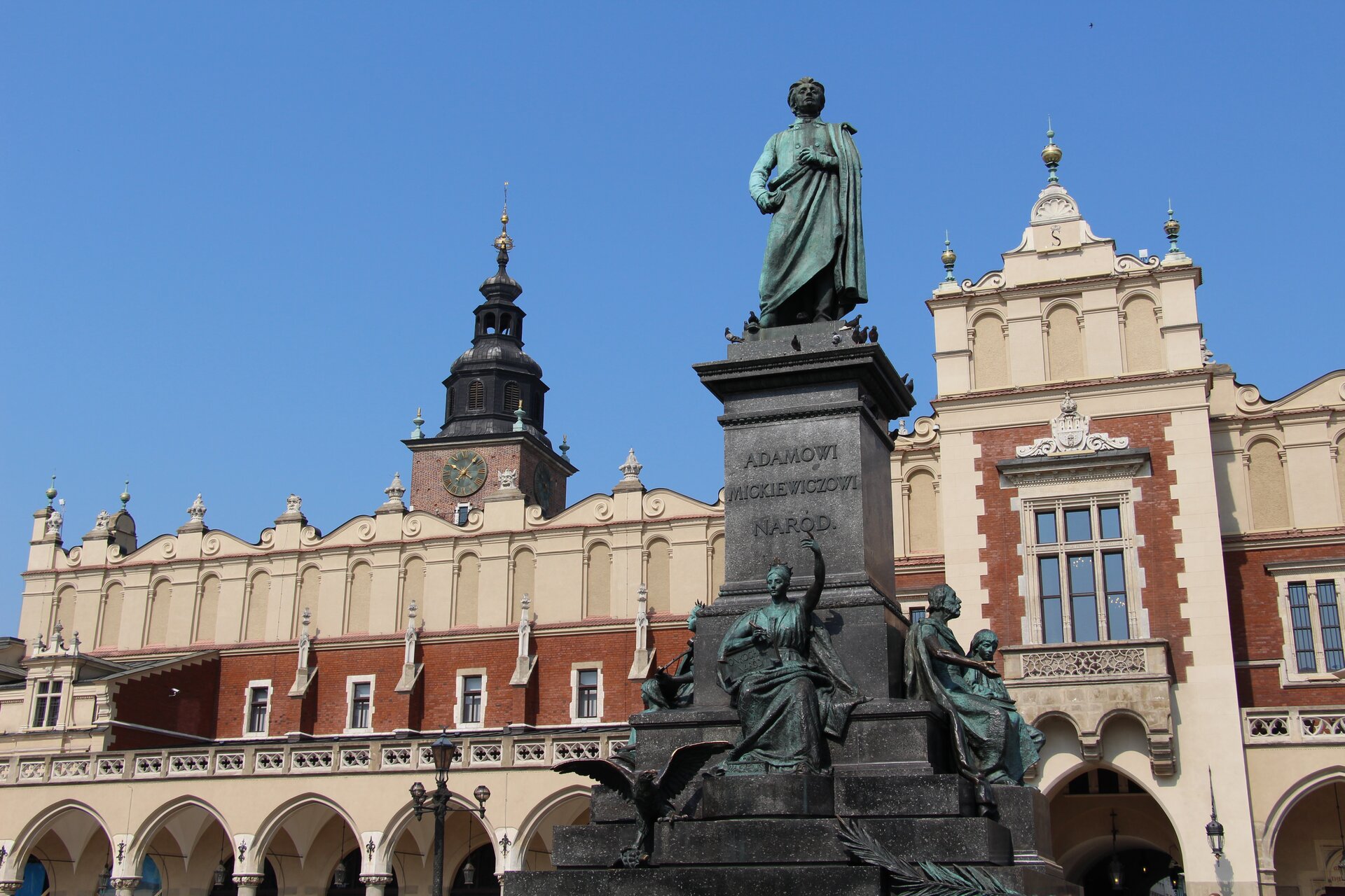 Zdjęcie przedstawia na tle błękitnego nieba oraz starego zadbanego budynku posąg mężczyzny w zielonkawym odcieniu, odzianego w luźno oplatające go szaty, ma lekko ugięte ręce, dumnie spogląda przed siebie. Poniżej znajdują się w nieco mniejszych rozmiarach posągi siedzących kobiet z wyciągniętymi przed siebie rękoma.