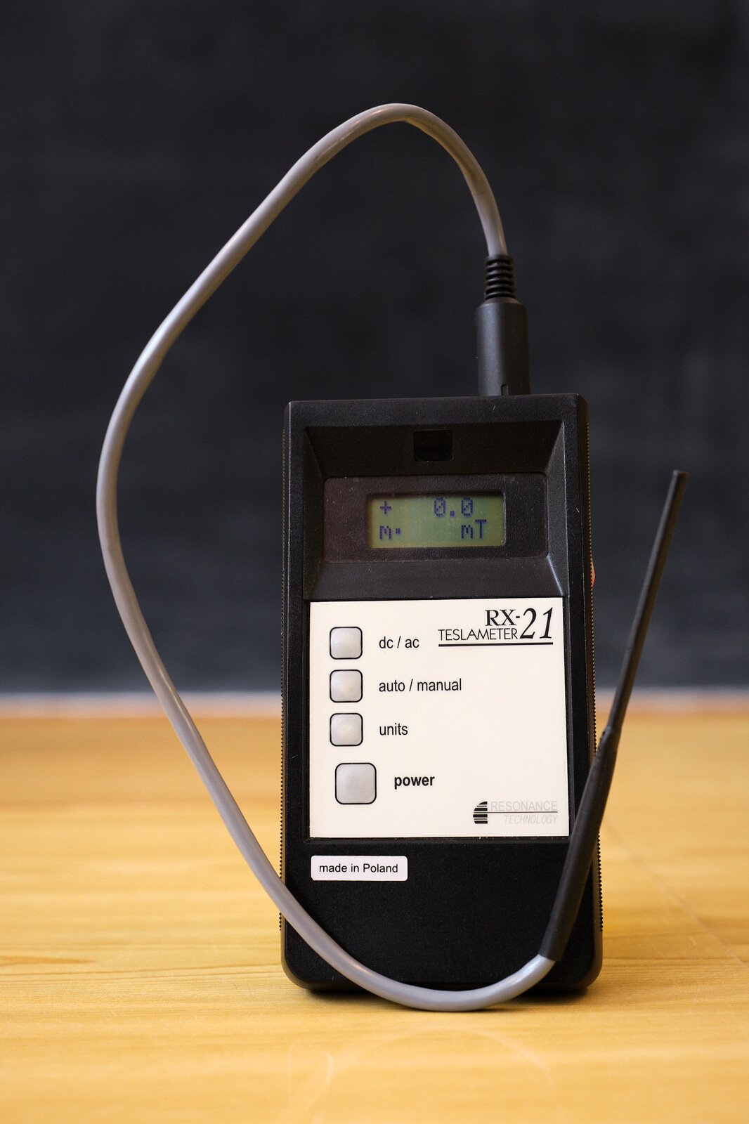 Rys. 2. Zdjęcie poglądowe przedstawia teslomierz. Teslomierz jest urządzeniem służącym do pomiaru wektora indukcji pola magnetycznego. W rzeczywistości jest to niewielki miernik z wychodzącą z jego górnej części sondą. Dokładniej do górnej części urządzenia przytwierdzony jest szary przewód, który biegnie w lewą stronę i w dół. Na końcu przewodu po prawej stronie ilustracji widoczny jest próbnik w postaci cylindrycznego, czarnego kształtu przypominającego długopis. W próbniku znajduje się cienka blaszka, która umieszczona w polu magnetycznym ulega odchyleniu, a odchylenie to jest proporcjonalne do wartości wektora indukcji magnetycznej, co umożliwia pomiar. Na ilustracji przedstawiono prostopadłościenne urządzenia z niewielkim ciekłokrystalicznym wyświetlaczem oraz białym panelem przednim. Na panelu umieszczone są przyciski dc/ac poniżej auto/manual poniżej units poniżej power. Widoczna jest również nazwa i model urządzenia teslameter RX‑21 oraz napis made in Poland.