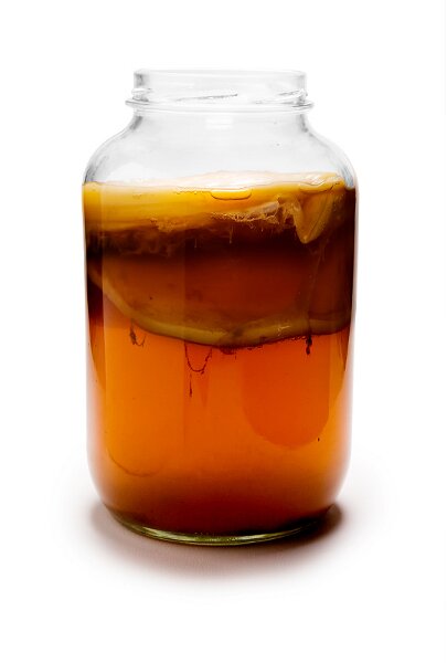 Zdjęcie szklanej butelki z kombuczanem. Płyn ma barwę brązową, półprzezroczystą. Na powierzchni znajduje się kożuszek różnych gatunków fermentujących bakterii. 