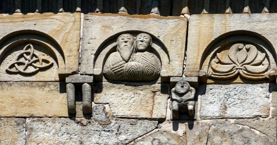 Fragment późnoromańskiego fryzu kamiennego na elewacji kościoła: reliefy z motywami ornamentalnymi i figuralnymi, w tym dwie uproszczone postacie ludzkie ujęte w półkolistej arkadce oraz drobne wsporniki pod gzymsem.