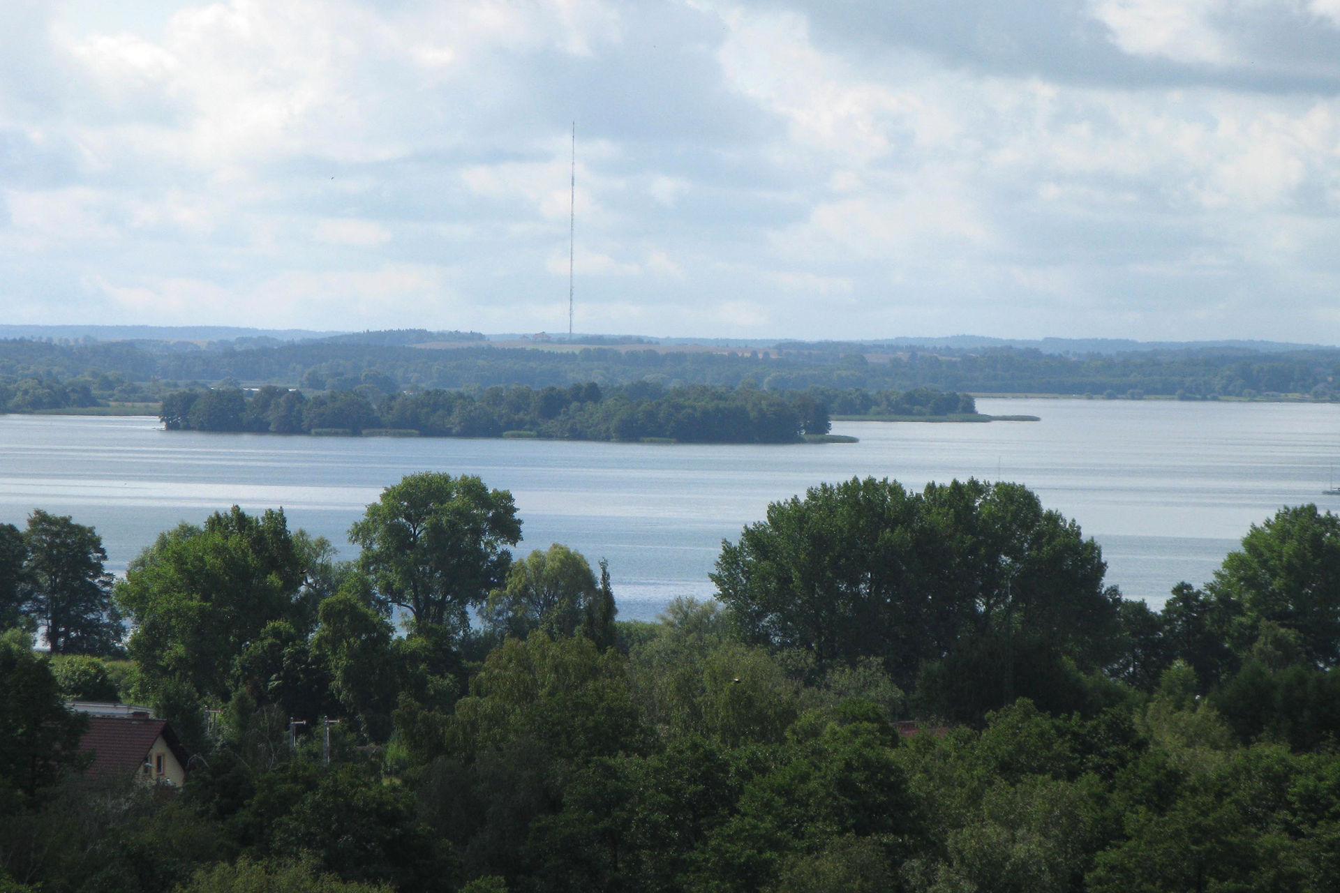 Fotografia prezentuje rozległe polodowcowe jezioro Niegocin, powstałe na skutek wędrówki olbrzymiego lodowca. Na jeziorze znajdują się wyspy porośnięte drzewami liściastymi. Jezioro z przodu i od tyłu również otaczają lasy.  Po prawej oraz po lewej stronie nie widać końca jeziora.