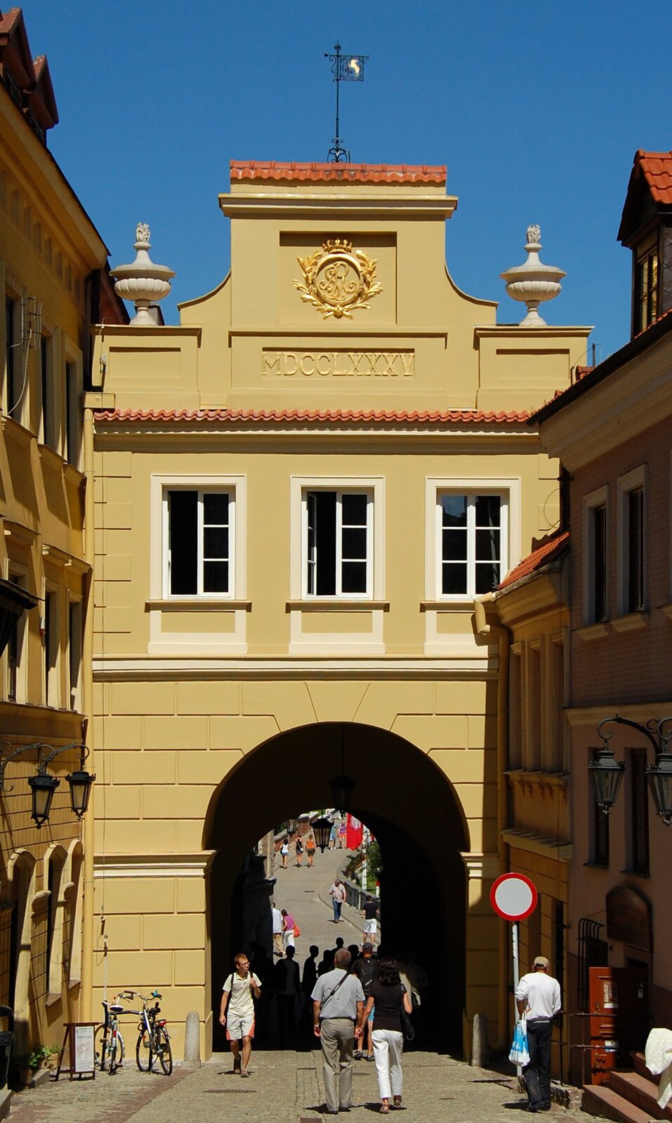 Zdjęcie przedstawia wąską ulicę. Pośrodku stoi brama miejska. Nad nią znajdują się trzy okna. Budowla jest zwieńczona inicjałem okolonym gałązkami. Pod nim umieszczono napis: MDCCLXXXV. Pod bramą przechodzą ludzie. Wzdłuż ulicy stoją kilkukondygnacyjne kamienice. W tle znajduje się bezchmurne niebo.