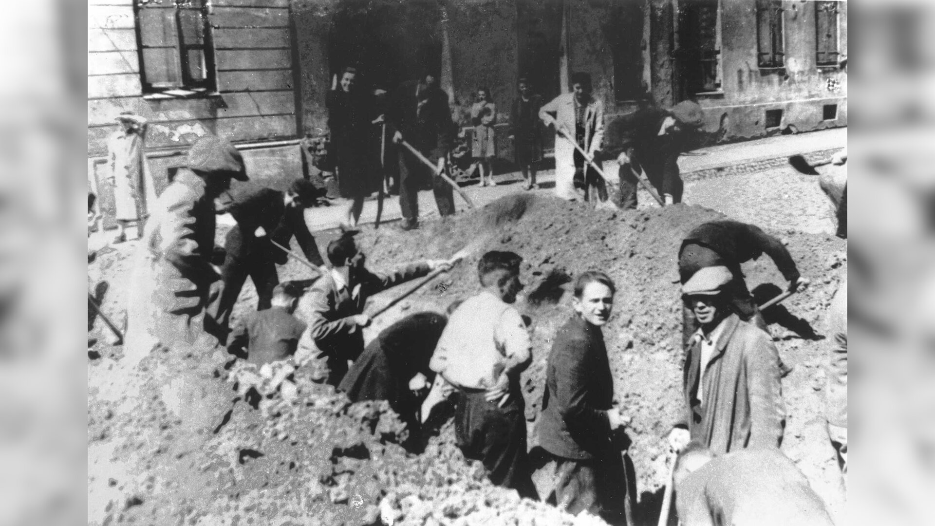 Zdjęcie przedstawia ulicę miasta, na której usypywane są barykady. Młodzi mężczyźni z łopatami w dłoniach wykonują przekop przez środek ulicy. W tle znajduje się kamienica.