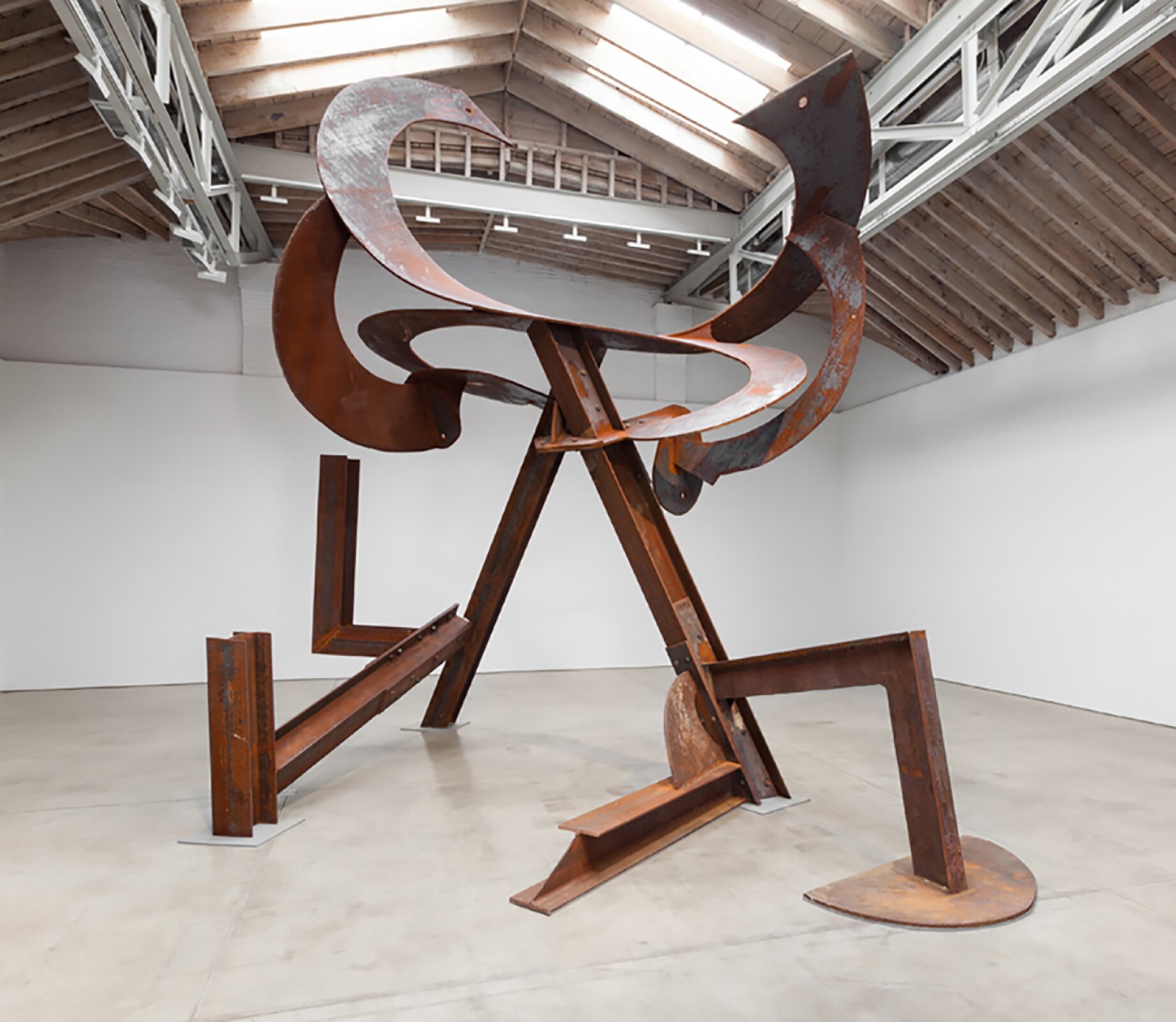 Mark di Suvero, widok instalacji „Mały tancerz” Fotografia przedstawia instalację umieszczoną w jasnym pomieszczeniu o wysokim suficie. Dzieło składa się z połączonych ze sobą metalowych elementów, przypominających miedziane bądź pokryte rdzą. Elementy przypominają grube blaszki, połączone zawiasami. Znajdują się części pionowe i poziome, wspierające konstrukcję. Przeważają figury geometryczne – prostopadłościany i półokręgi. U góry umieszczono kilka elementów łukowych, swoim kształtem przypominające sierpy.