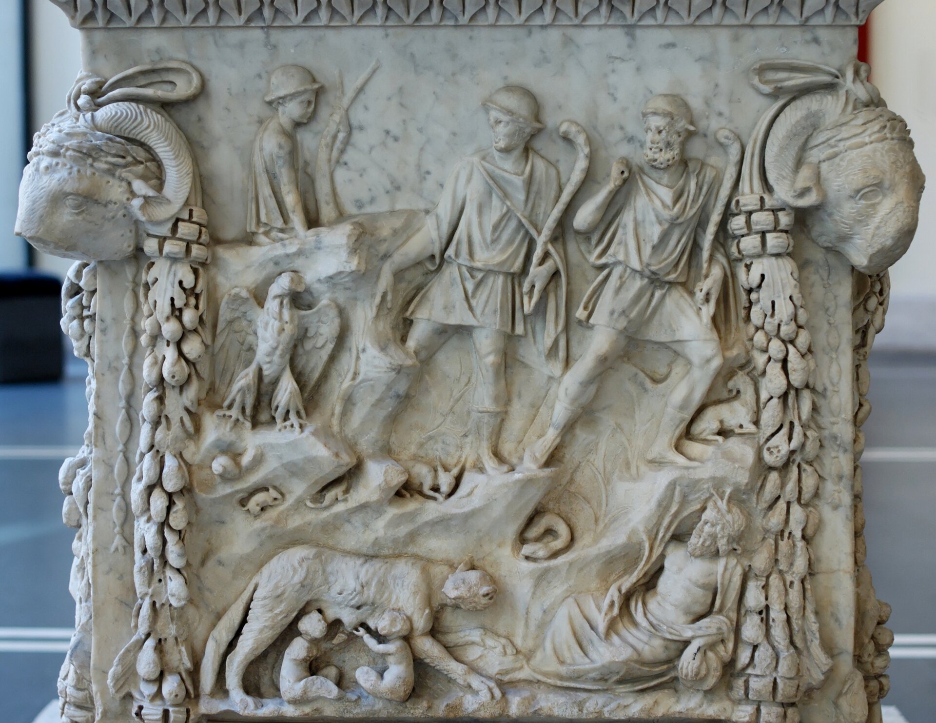 Ilustracja przedstawia relief obrazujący mit założenia Rzymu. Na reliefie widzimy Romulusa i Remusa karmionych przez wilczycę. Nad wilczycą pojawia się również dwóch mężczyzn w krótkich tunikach oraz po lewej stronie żołnierz z bronią. Pod żołnierzem znajduje się ptak z dużymi rozłożonymi skrzydłami. 