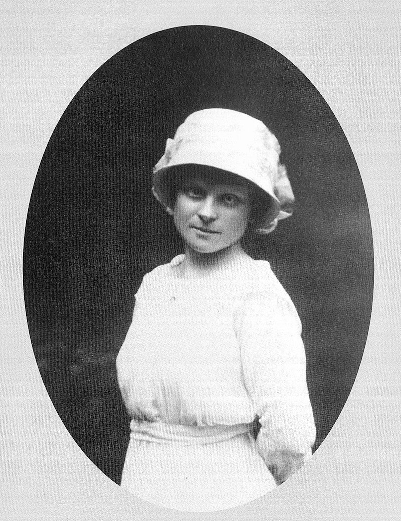 Zdjęcie przedstawia portret młodej kobiety. Jest szczupła. Jej twarz ma regularne rysy. Kobieta ma złożone usta w półuśmiechu. Patrzy na wprost siebie. Na głowie ma głęboki kapelusz z małym rondem. Ubrana jest w prostą, białą suknię. Trzyma ręce za plecami.