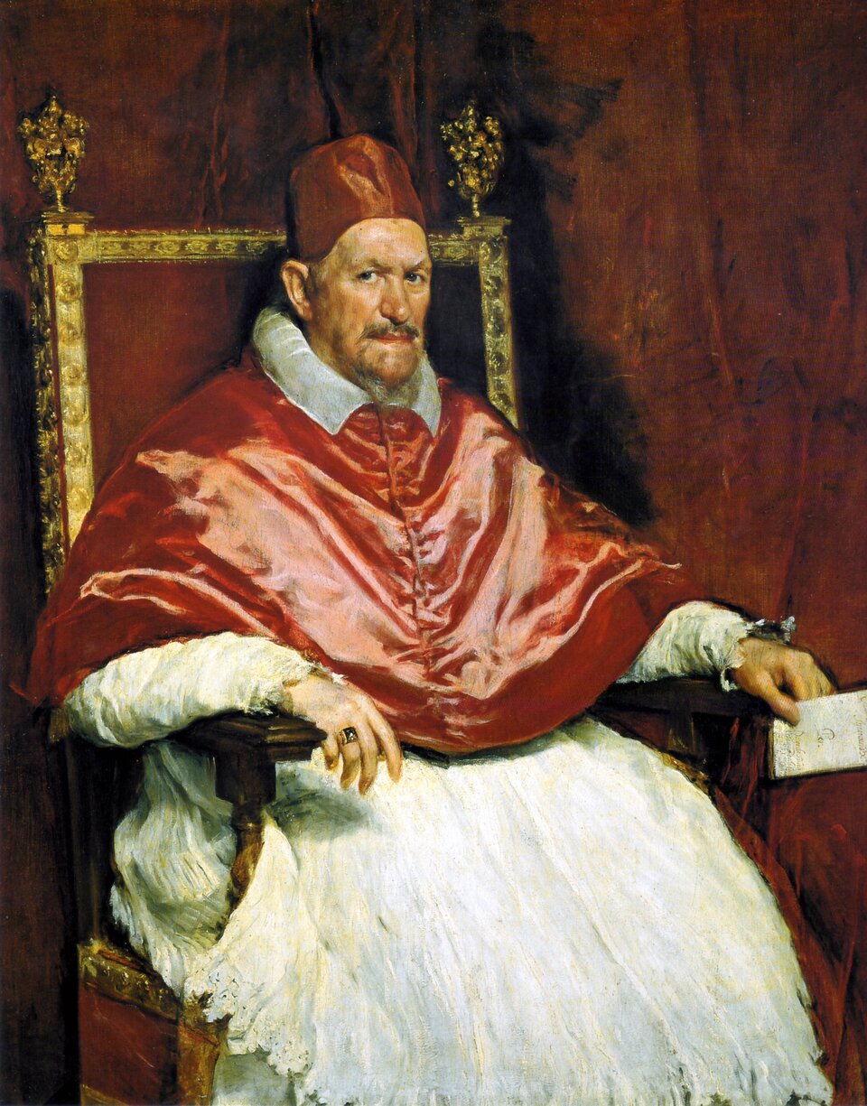 Ilustracja przedstawia „Portret Innocentego X” Diego Velázqueza. Mężczyzna siedzi na eleganckim fotelu, ukazany lekko z ukosa. Spoczywa z rękami ułożonymi na oparciach fotela i patrzy bezpośrednio na odbiorcę. Ma na sobie białe, obszerne, zwiewne szaty oraz krótką, czerwoną pelerynę zapinaną na guziki. Na głowie nosi nakrycie wykonane z tego samego czerwonego materiału. Na jego twarzy widoczne są zmarszczki mimiczne; ma zarost i zmarszczone brwi. Fotel, przypominający tron, ma ciemnoczerwony kolor i złote wykończenia na krawędziach. Tło obrazu jest czerwone.
