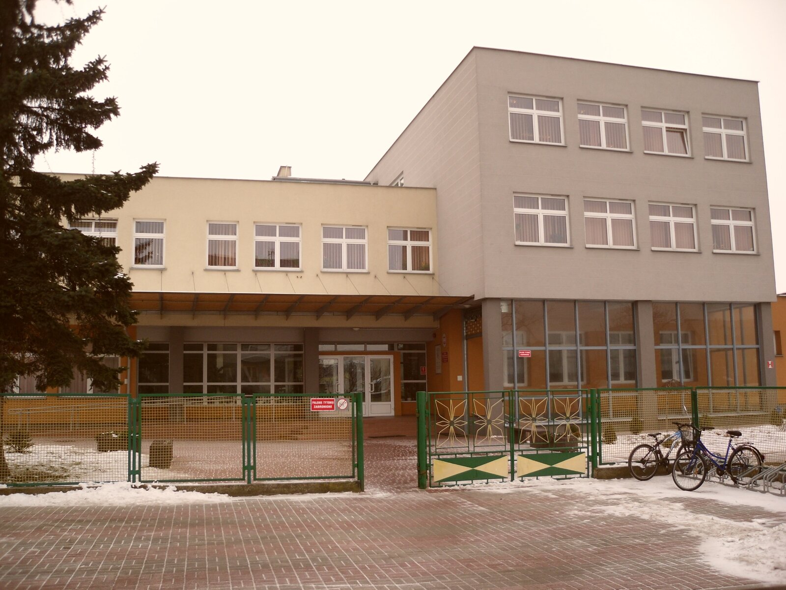 Ilustracja przedstawia budynek trzykondygnacyjny w kolorze szarym. Drzwi i okna w kolorze białym. Plac przed budynkiem wyłożony jest kostką brukową. Budynek ogrodzony jest metalowym płotem wykonanym z siatki w kolorze zielonym.