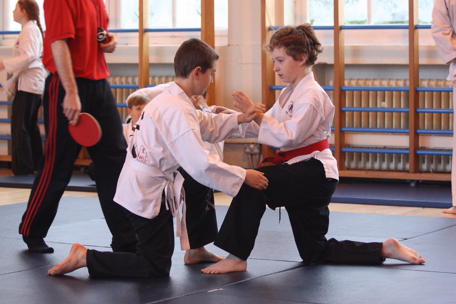 Na zdjęciu widoczna jest para dzieci, które trenują sztuki walki na specjalnej macie. Ubrane są w tradycyjne białe kimona i czarne spodnie. Znajdują się w pozycji klęcznej, dotykają się dłońmi. W tle można dostrzec drabinki, tworzące atmosferę sali treningowej.