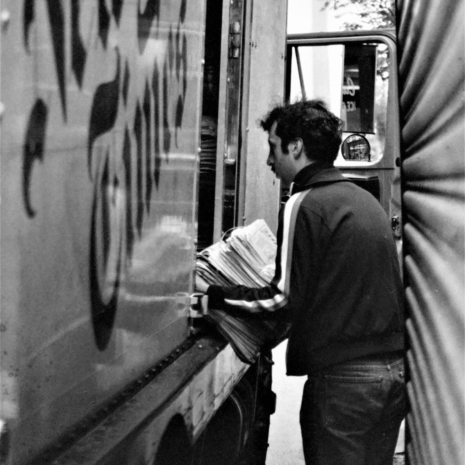 Czarno‑białe zdjęcie przedstawia stojącego przy ciężarówce mężczyznę, pokazanego do wysokości kolan. Mężczyzna ubrany jest w bluzę i spodnie, ma krótkie czarne włosy. Mężczyzna zwrócony jest w lewo i na ugiętych rękach ma stos gazet, które wyjmuje z przyczepy ciężarówki. Drzwi od strony pasażera są otwarte. Po prawej stronie widoczna jest zasunięta metalowa roleta.