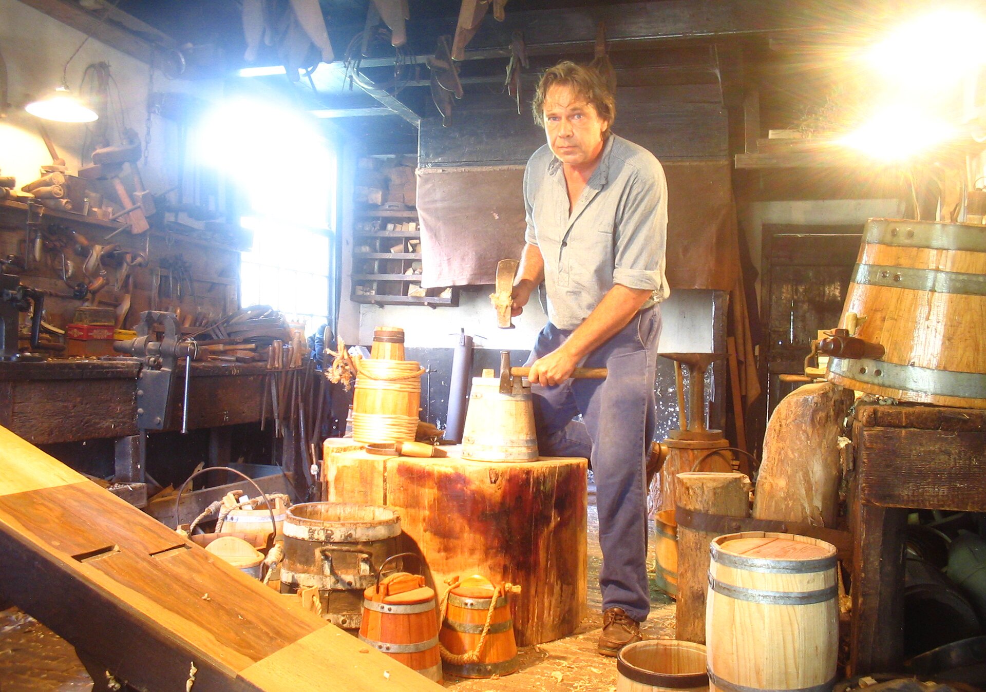 Fotografia przedstawia mężczyznę w warsztacie rzemieślniczym. W rękach trzyma młotki. Jeden z nich opiera o stojącą przed nim na pniu drzewa małą beczkę. Ubrany jest w roboczą koszulę i spodnie. Patrzy na wprost. Otaczają go rozmieszczone w różnych częściach pracowni beczki. Z lewej strony widoczny jest stół, na którym są narzędzia, m.in. imadło, hebel, lampa oświetlająca stół.