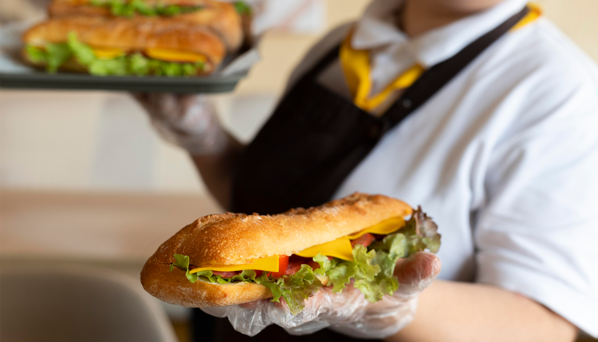 Zdjęcie przedstawia osobę, która trzyma na dłoni kanapkę. Widoczna jest sałata, ser i plastry pomidora. 