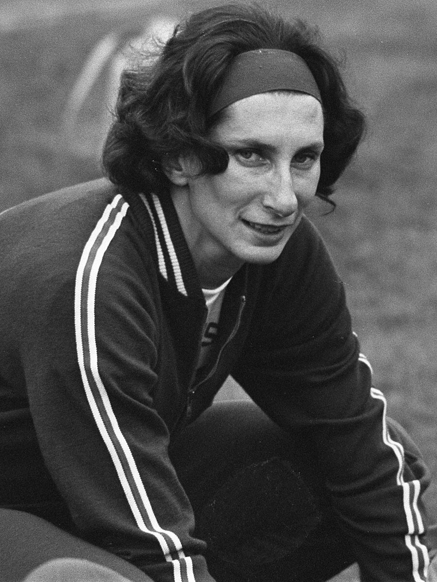 Zdjęcie przedstawia kucającą Irenę Szewińską. Kobieta ma sięgające ramion ciemne włosy. Ubrana jest w dres.