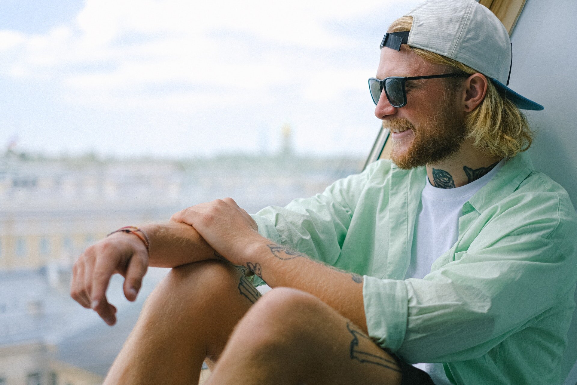 Zdjęcie przedstawia młodego, jasnowłosego mężczyznę. siedzącego na parapecie okna. Mężczyzna ubrany jest w podkoszulek, koszulę i krótkie szorty, na głowie ma czapkę bejsbolówkę. Ma założone okulary przeciwsłoneczne.