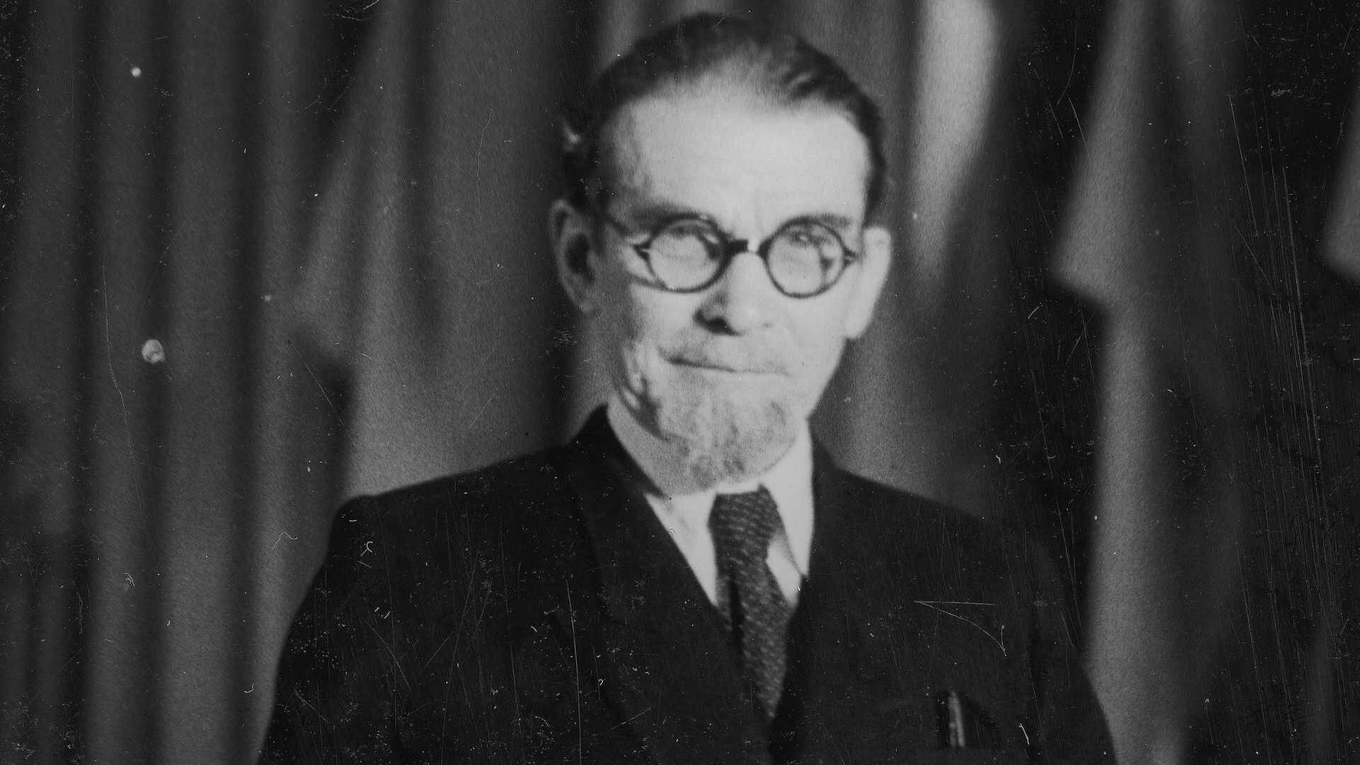 Fotografia portretowa mężczyzny w średnim wieku, ubranego w garnitur, koszulę i krawat. Mężczyzna ma krótkie włosy zaczesane do tyłu, wąsy, krótką brodę wokół ust i okrągłe okulary.