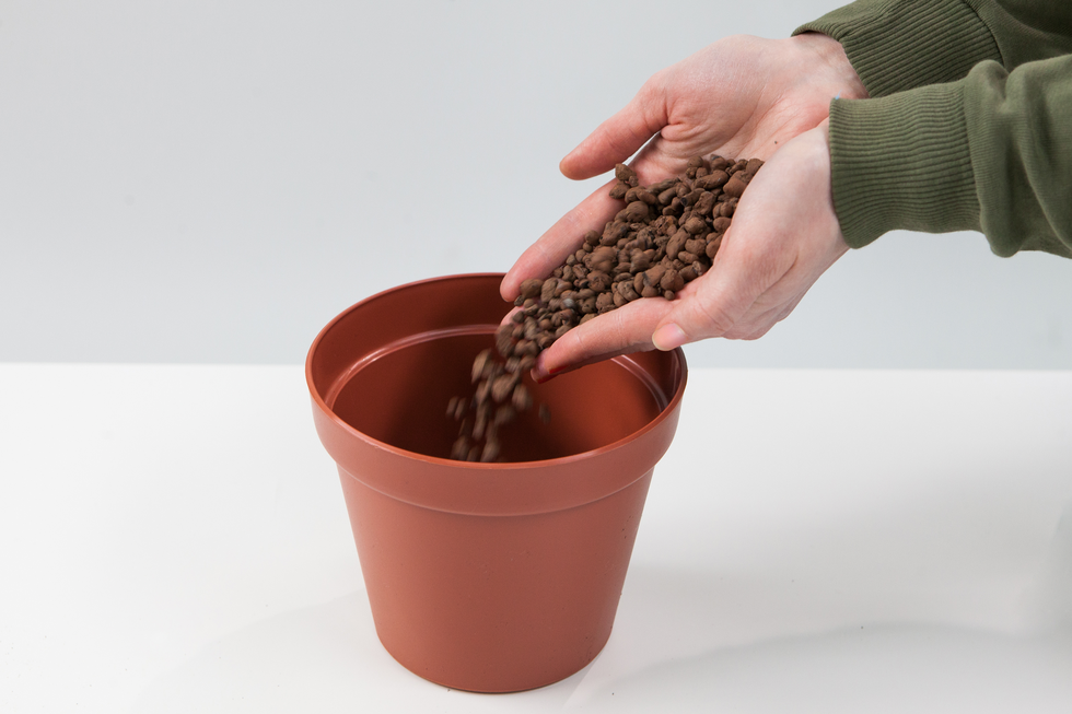 Fotografia przedstawia dłonie osoby, która nasypuje żwir do brązowej doniczki