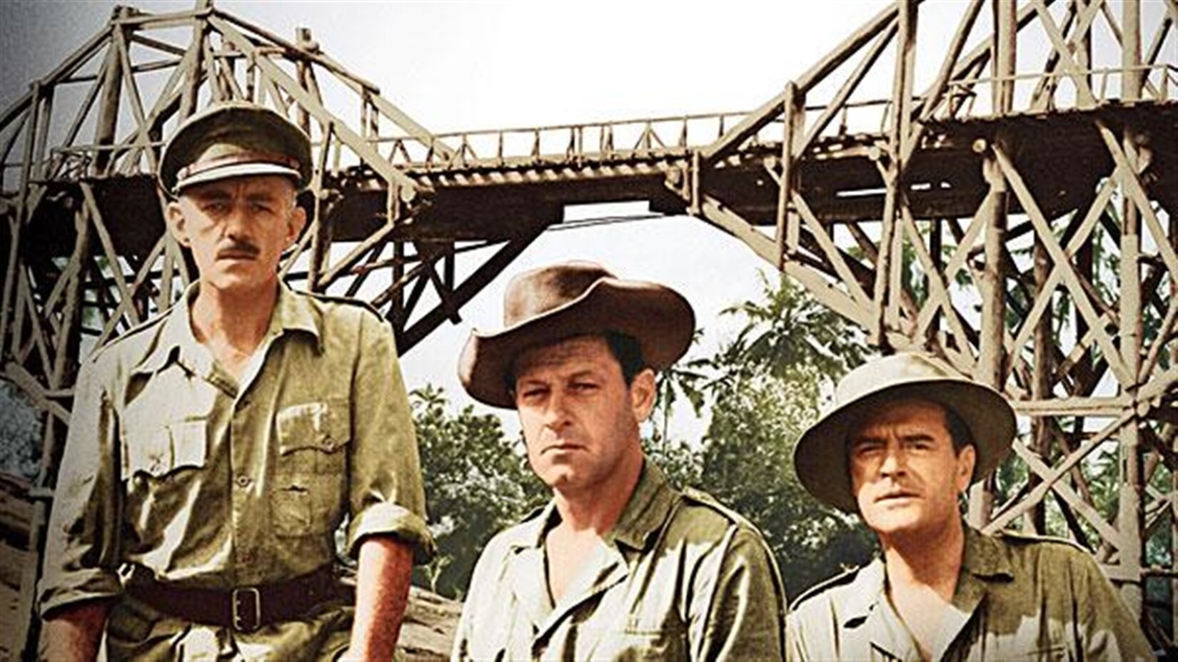 Kadr z filmu „Most na rzece Kwai” w reżyserii Davida Leana, przedstawiający trzech mężczyzn w koszulach koloru khaki oraz kapeluszach na tle budowanego mostu. Jeden z mężczyzn, uchwycony z lewej strony, stoi patrząc przed siebie. Widoczny jest od pasa w górę – w talii ma brązowy pas. Ma podkrążone oczy, czarne, krótkie wąsy oraz zmarszczki pomiędzy brwiami. Pozostali dwaj mężczyźni widoczni są od ramion w górę. Spod kapeluszy wystają fragmenty ich ciemnych włosów. Mężczyzna uchwycony w środku wydaje się młodszy od pozostałych, zmarszczki na jego twarzy są mniej wyraźne. Brwi mężczyzny siedzącego z prawej strony są zmarszczone, a usta lekko otwarte. W tle widać jasne niebo oraz zielone liście. 