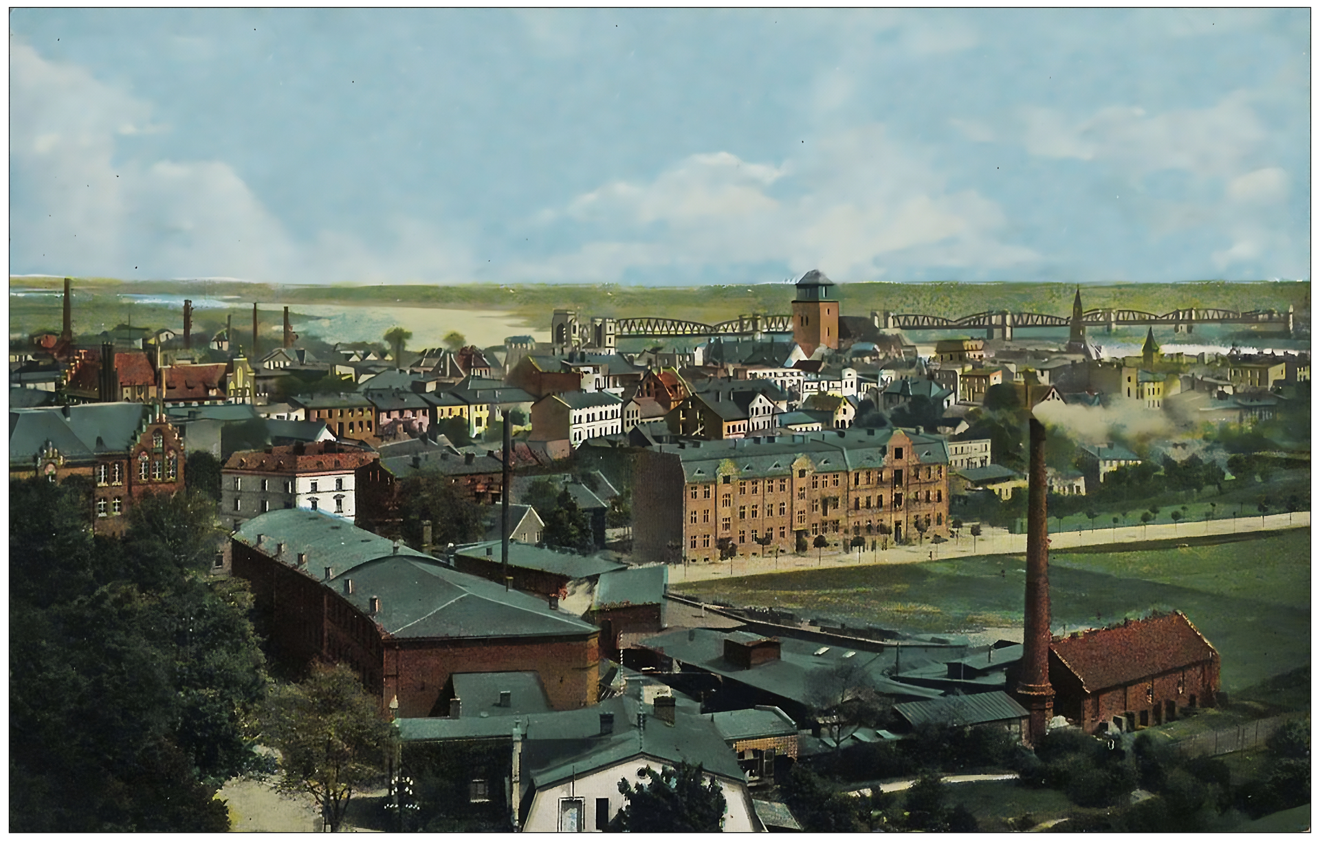 Zdjęcie przedstawia widok na miasto. W górnej części zdjęcia, a także po lewej stronie, widoczne są kilkupiętrowe, ceglane kamienice ze skośnymi, ciemnymi dachami. Zabudowania są gęsto ułożone, a pomiędzy nimi widoczne są wysokie kominy. Na pierwszym planie, u dołu zdjęcia, po prawej stronie znajduje się długi, ceglany budynek, z lekko skośnym dachem. Po prawej stronie znajduje się mały ceglany budynek do którego przylega wysoki komin, z którego wydostaje się dym. Obok, z prawej strony zdjęcia, duże, zielone pole. Za nim szeroki, trzypiętrowy budynek mieszkalny. Na dalszym planie kościół z wysoką wieżą. W tle miasta, za budynkami, w górnej części zdjęcia, widoczna jest rzeka i metalowy most łączący dwa brzegi. 