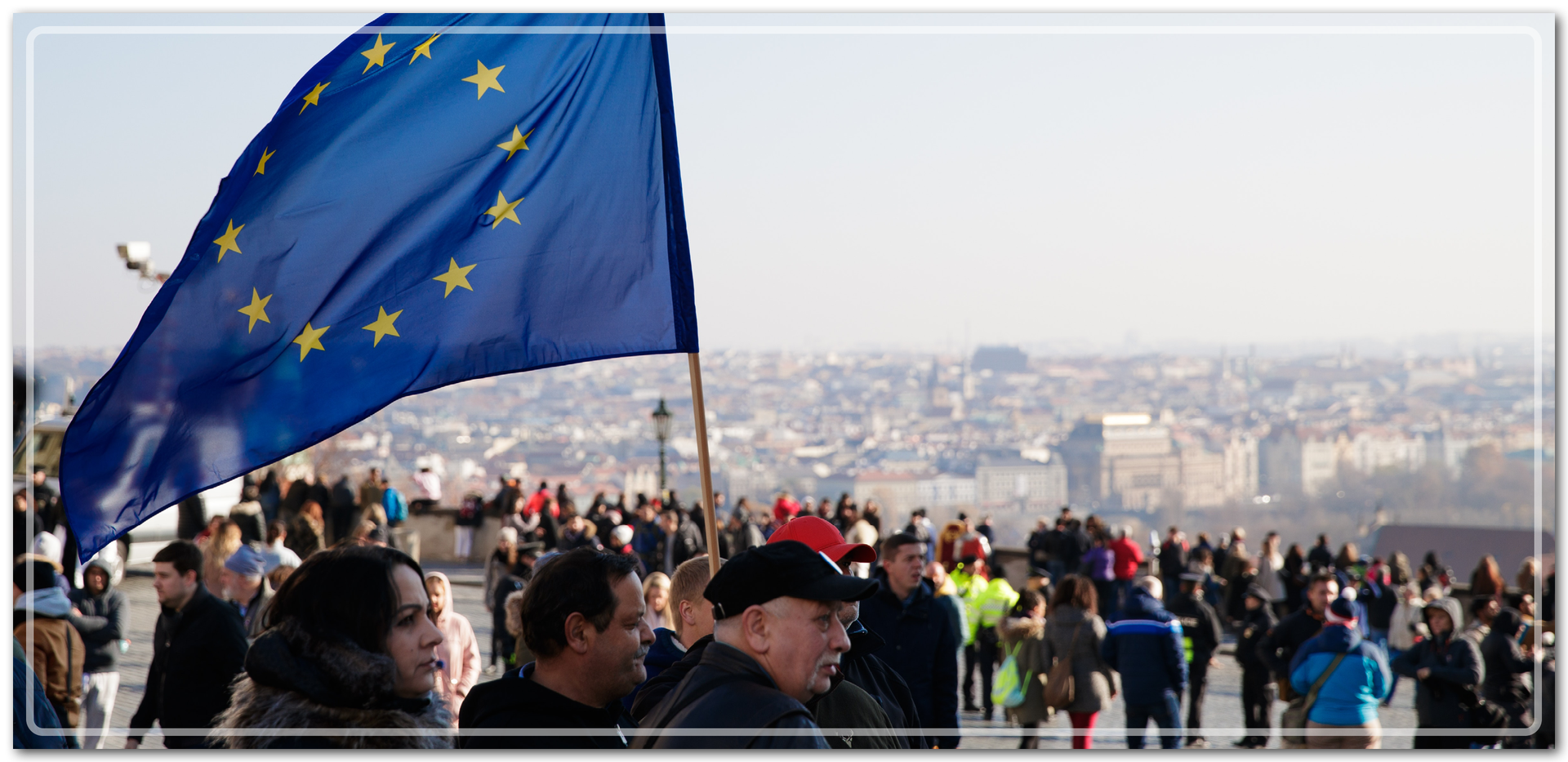 Ilustracja przedstawia tłum ludzi. Jeden z ludzi niesie flagę europejską, czyli niebieską flagę z okręgiem złożonym z dwunastu złotych gwiazd.