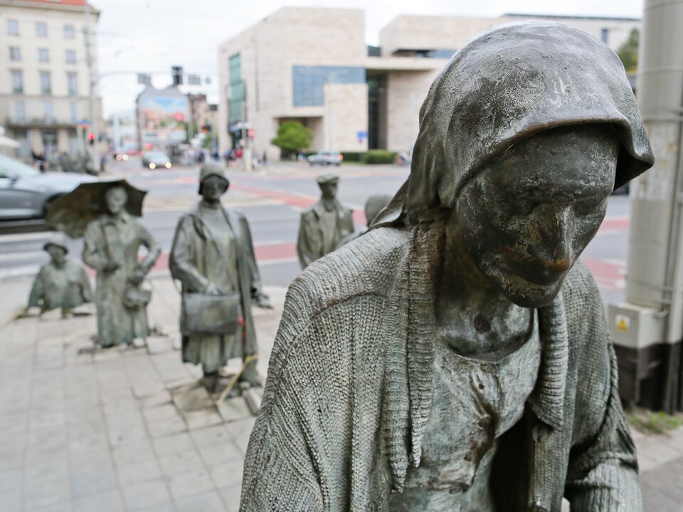 Zdjęcie przedstawia postaci odlane z brązu umieszczone w miejskiej przestrzeni - ulicznym chodniku. Przypominają one zastygłych w bezruchu przechodniów. Osoba na pierwszym planie to starsza kobieta. Ma opuszczoną głowę, na głowie chustkę. Ubrana jest w naciągnięty sweter. Za nią znajduje się kobieta w kapeluszu z torbą trzymaną w prawej dłoni. Po prawej stronie kobiety postawiono odlaną z brązu figurę mężczyzny. Za kobietą kolejna postać — kobieta z rozłożoną parasolką, następna osoba ukazana jest tylko do połowy, wygląda, jakby zapadła się w głąb ulicznego chodnika. Im bliżej posągi osób znajdują się ulicznej krawędzi widocznej w tle, tym głębiej zapadają się w chodnikowych płytach. W tle ulica miasta z samochodami i budynkami.