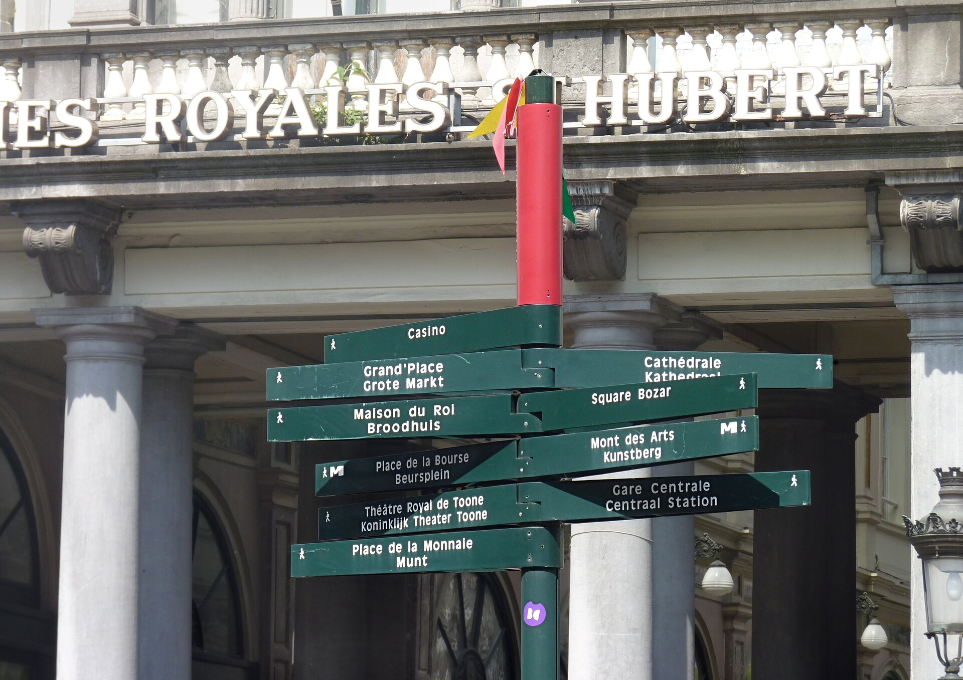 Na zdjęciu zielony słupek z licznymi drogowskazami. Na zielonym tle białe napisy w dwóch językach, francuskim i flamandzkim. W tle budynek.