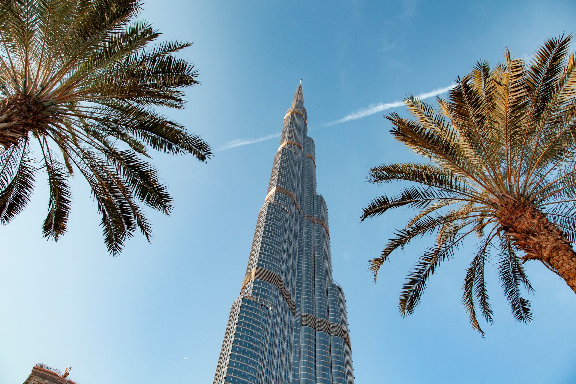 Ilustracja przedstawia Burdż Chalifa - najwyższy budynek na świecie, liczący 828 metrów wysokości. Na tle błękitnego nieba – patrząc od dołu - widać wysoką budowlę, a obok niej dwie palmy. 