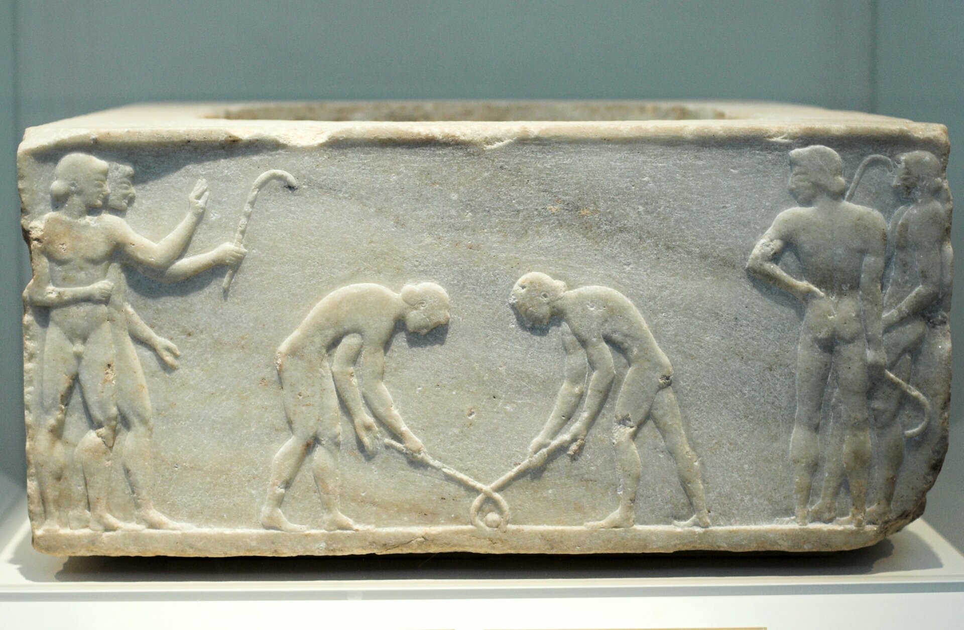 Ilustracja przedstawia płaskorzeźbę. Są na niej nadzy mężczyźni. Trzymają zakrzywione kije. W centralnym miejscu płaskorzeźby dwóch mężczyzn trzyma kije ,pomiędzy którymi jest mała kula.  