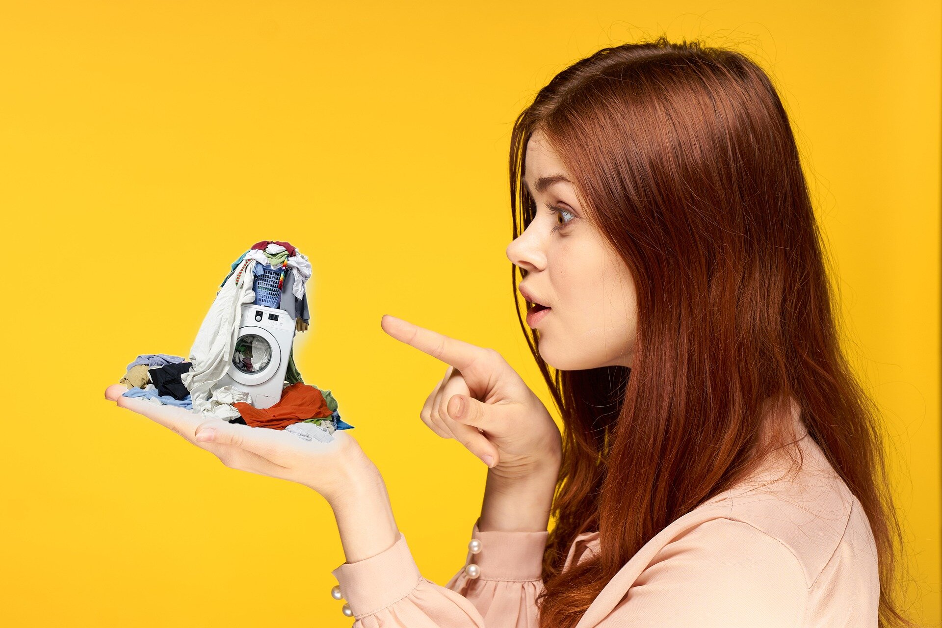 Na zdjęciu obserwujemy młodą kobietę, która wskazuje na swoją dłoń, na której stoi miniaturowa pralka. Na pralce znajduje się kosz z odzieżą, która jest także porozrzucana wokół niej.