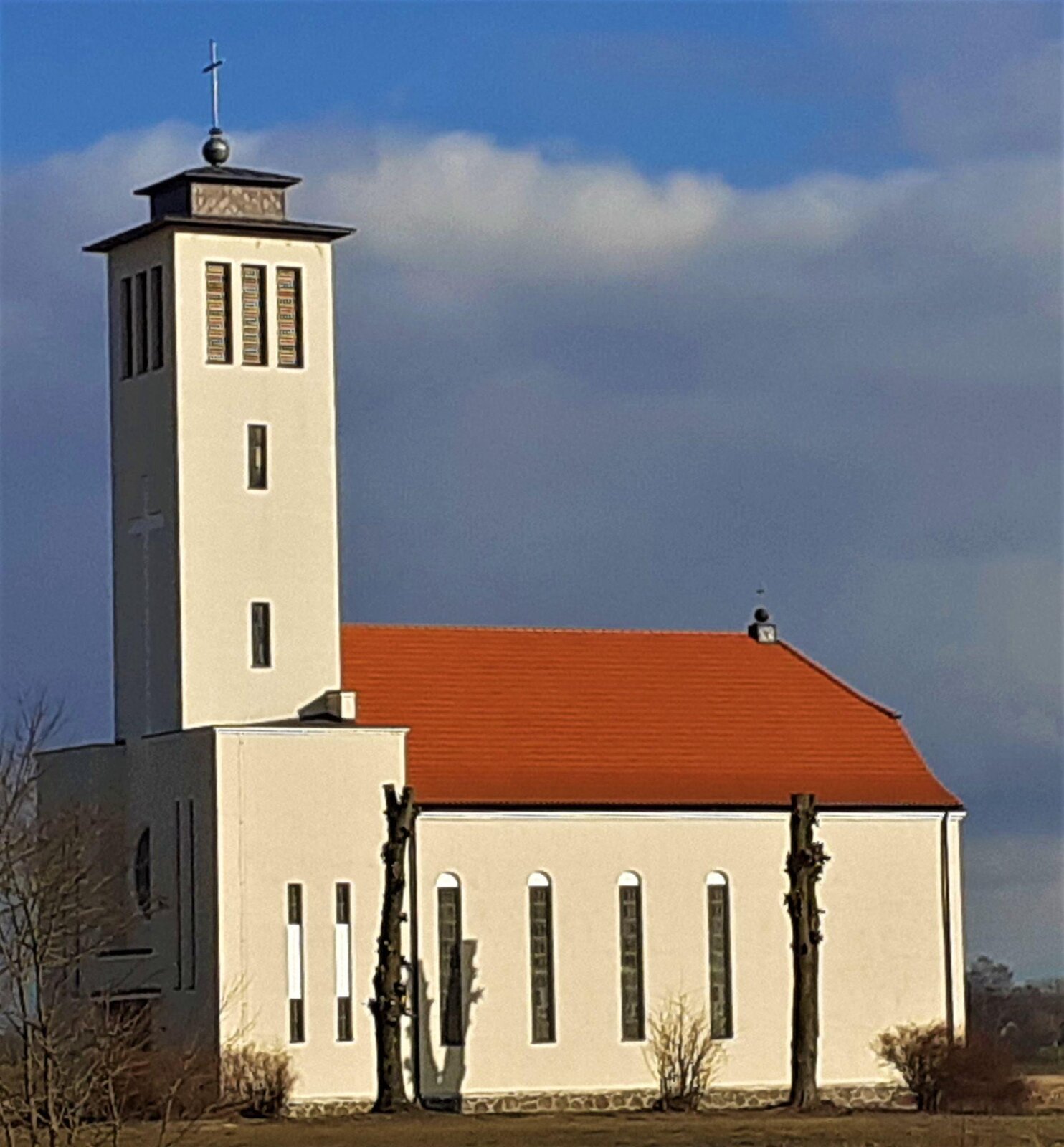 Zdjęcie przedstawia kremowy kościół z pomarańczowym dachem. Z przodu budynku znajduje się wysoka wieża, której czubek zwieńczony jest krzyżem. Okna są podłużne, wysokie i wąskie. Wokół kościoła widoczne są fragmenty bezlistnych drzew. Niebo jest błękitne, częściowo zasłonione białymi chmurami. Zdjęcie zostało wykonane w świetle dziennym.