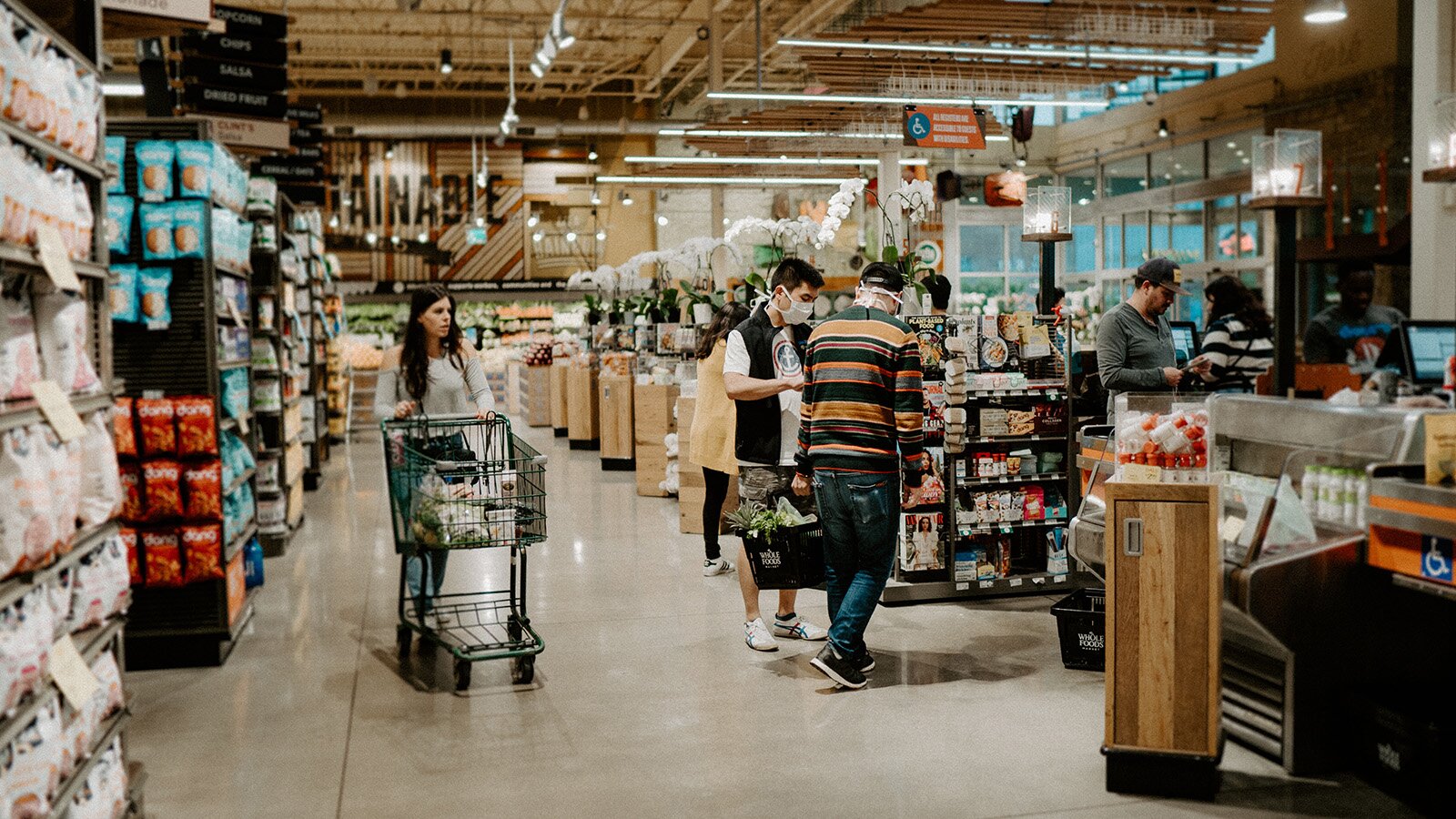 Zdjęcie ukazuje koniec alejek sklepowych w supermarkecie, w miejscu gdzie zaczynają się kasy. W kierunku kas zmierza dwóch mężczyzn niosących koszyk. Mężczyźni mają maseczki na twarzy. Za nimi idzie młoda kobieta prowadząca wózek wypełniony w połowie zakupami.